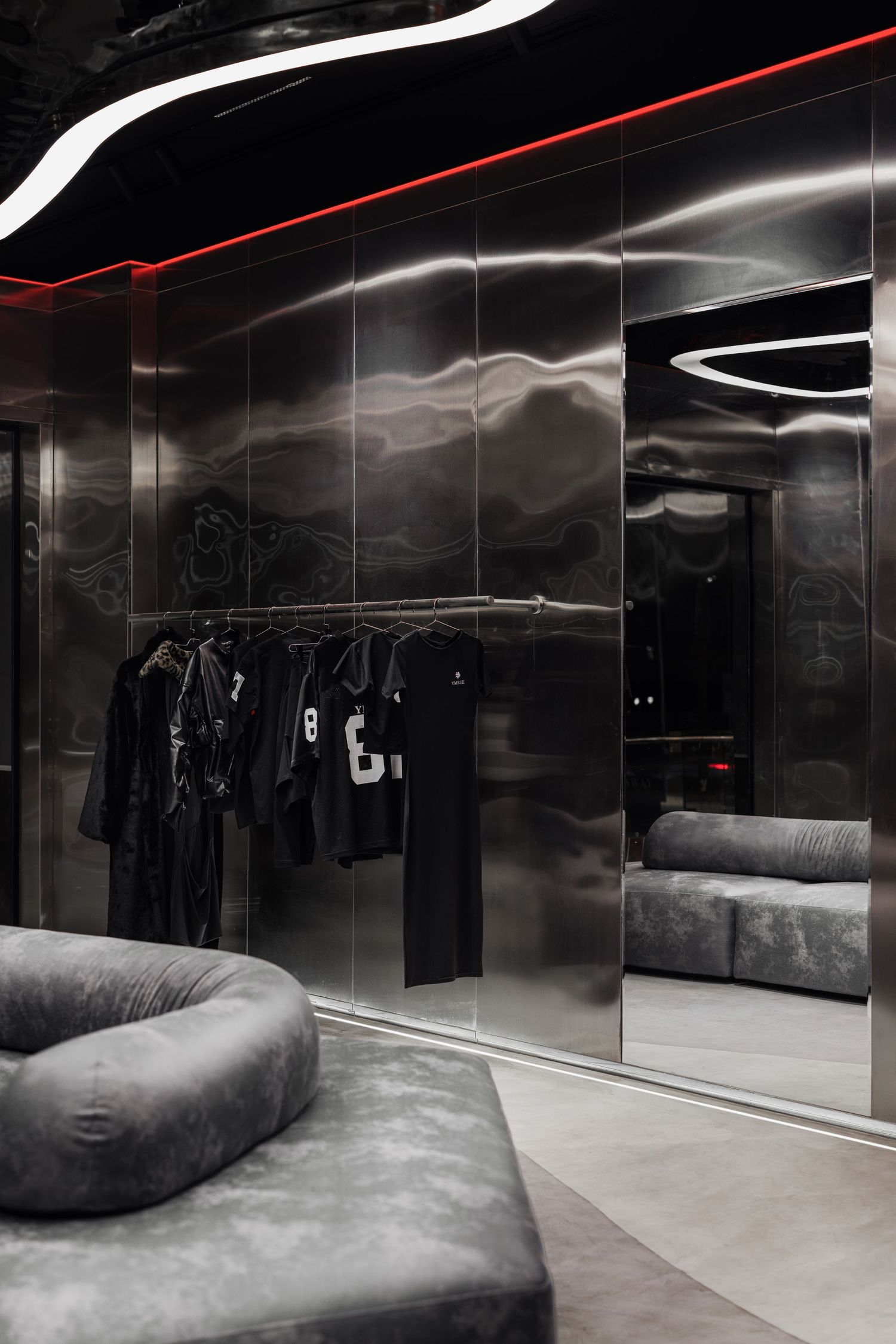 Dark luxury clothing store with metallic ripple walls, black garments on rack, gray velvet seating, and curved white LED lights.