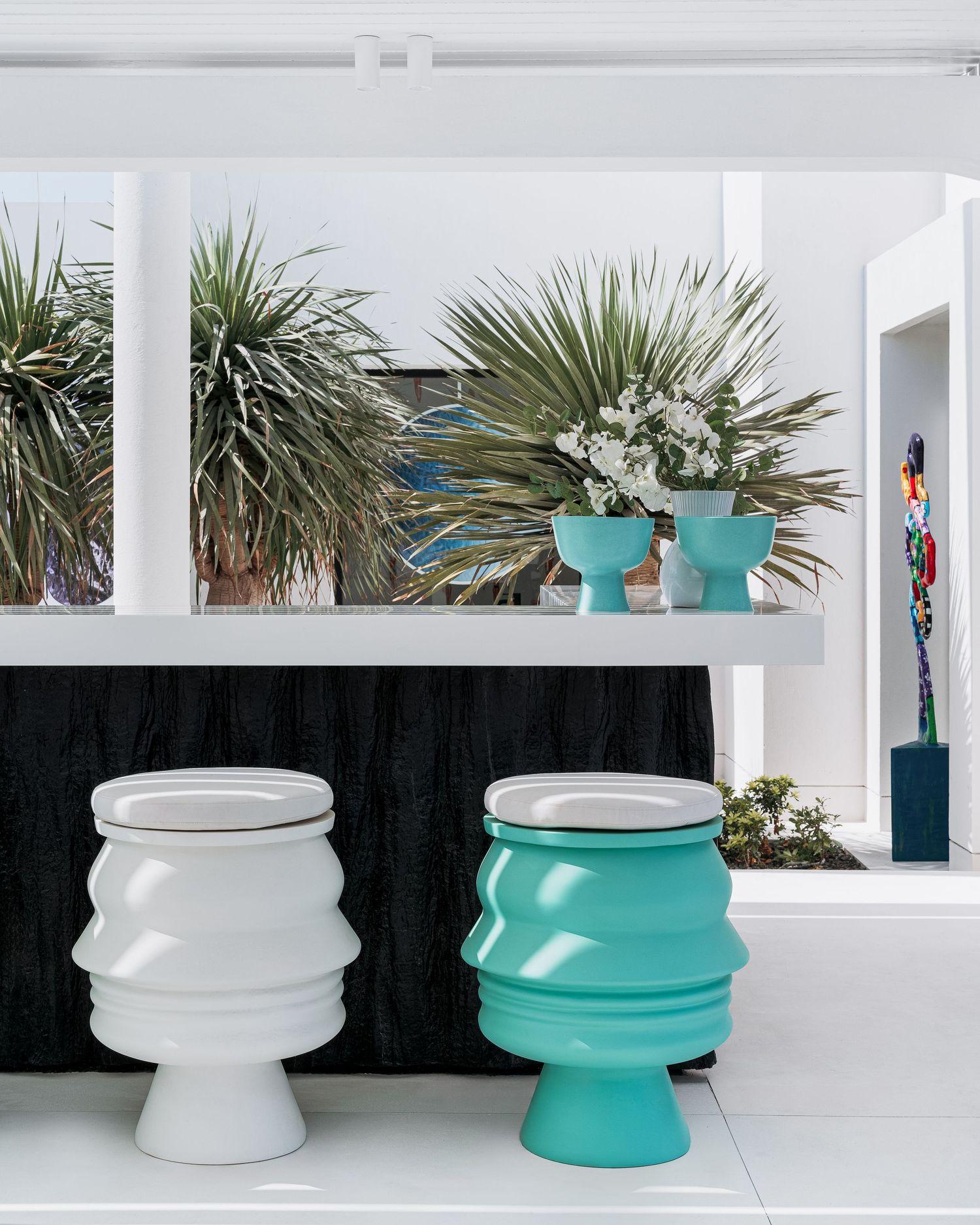 Two sculptural ribbed stools in white and turquoise displayed beside matching vases with tropical plants in a modern white interior.