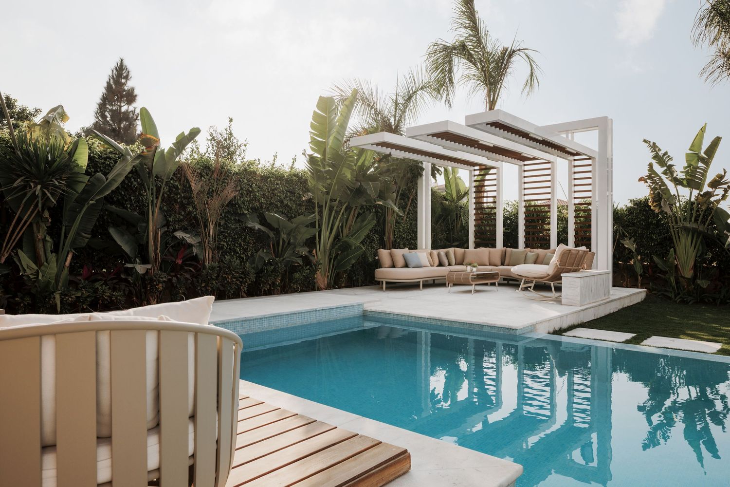 Luxury outdoor pool with white pergola, tropical plants, and beige lounge seating.