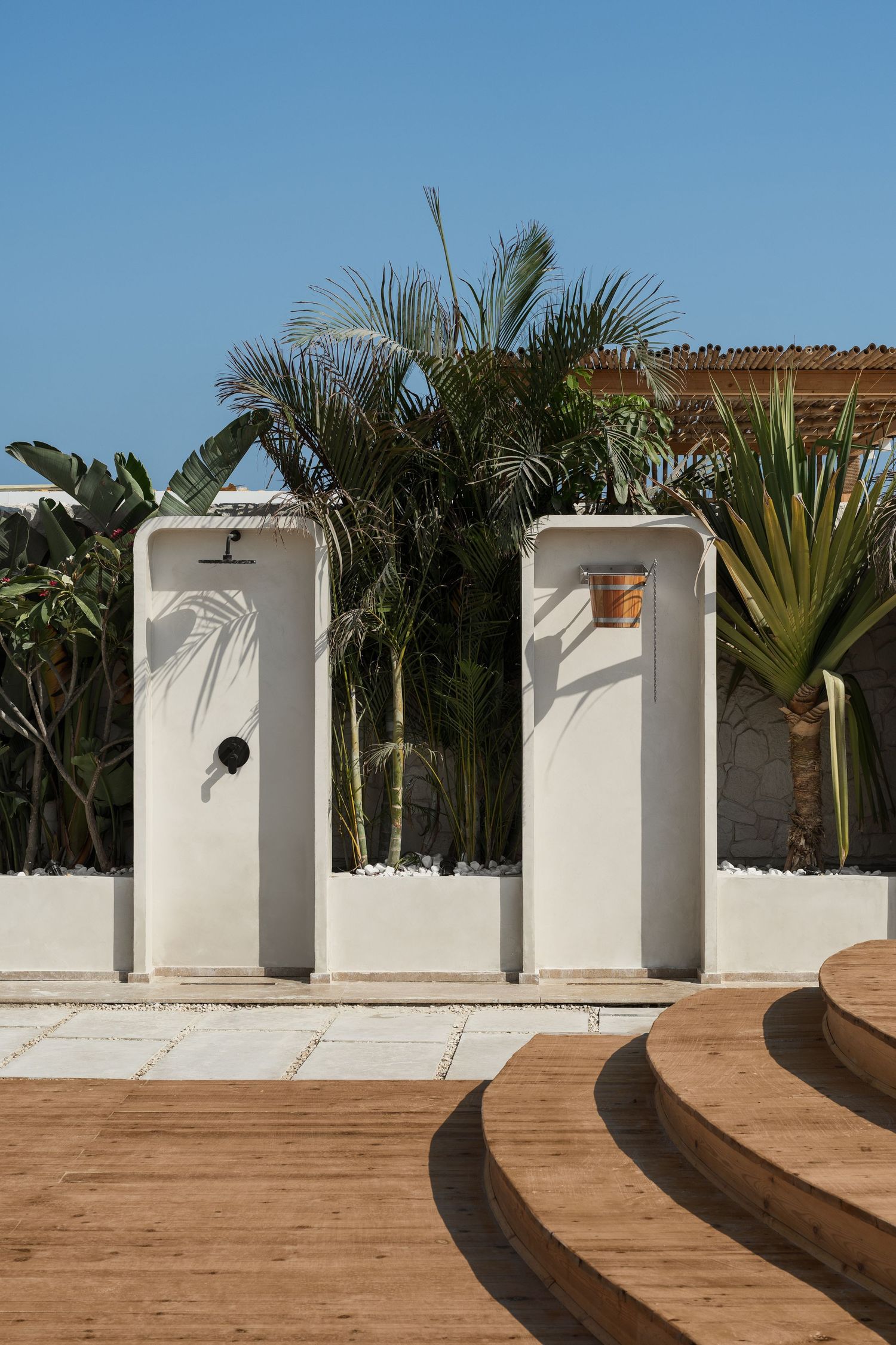 Two white outdoor shower stations with wooden buckets surrounded by lush tropical palm trees.