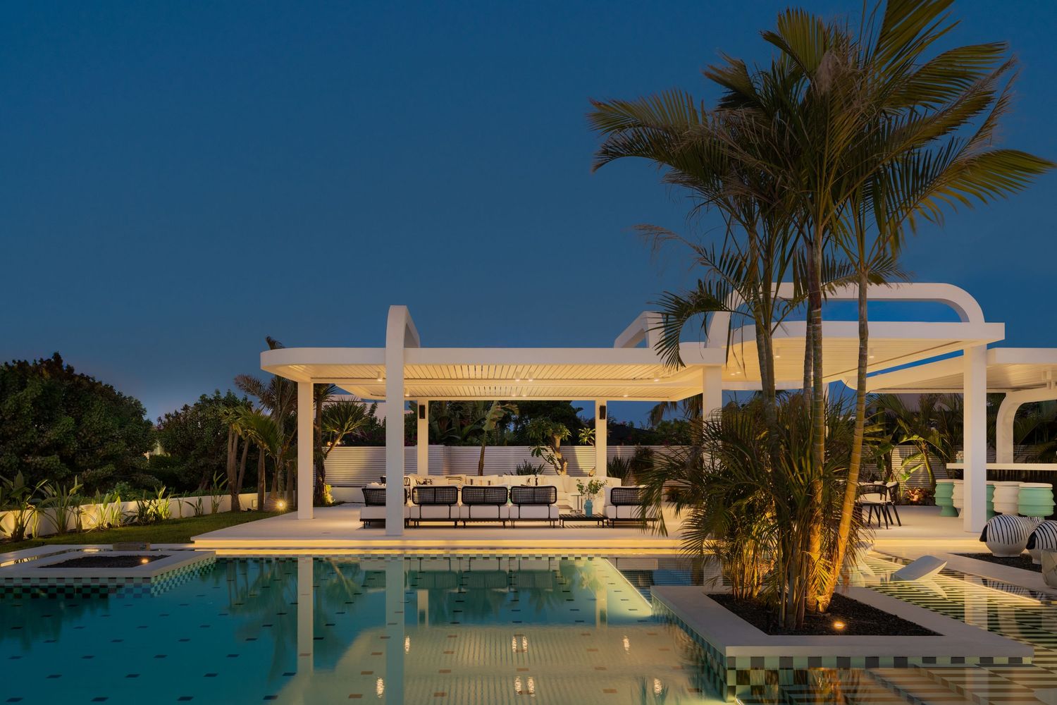 Elegant white pergola and outdoor lounge area beside a serene illuminated pool surrounded by tall palm trees at twilight.