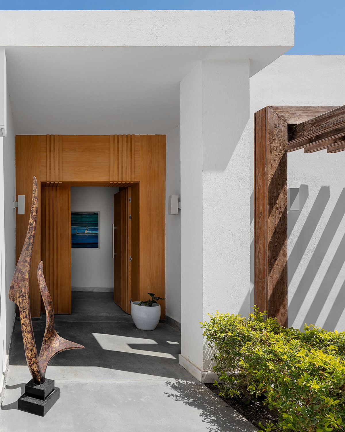 Modern home entrance with wooden door, abstract sculptures, white walls, and lush greenery under bright sunlight.