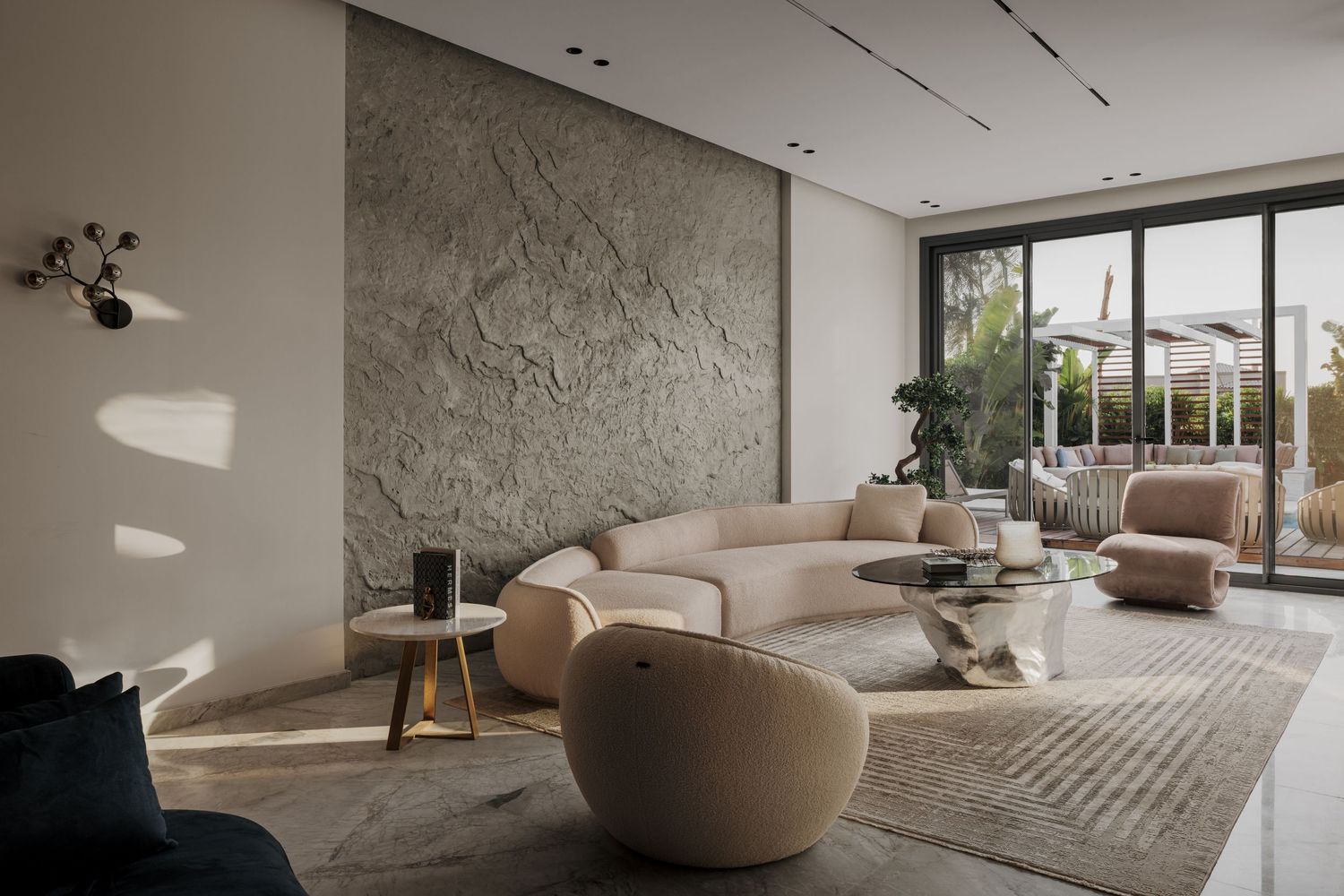 Bright modern living room with curved beige sofa, stone textured accent wall, glass coffee table, and large floor-to-ceiling windows.