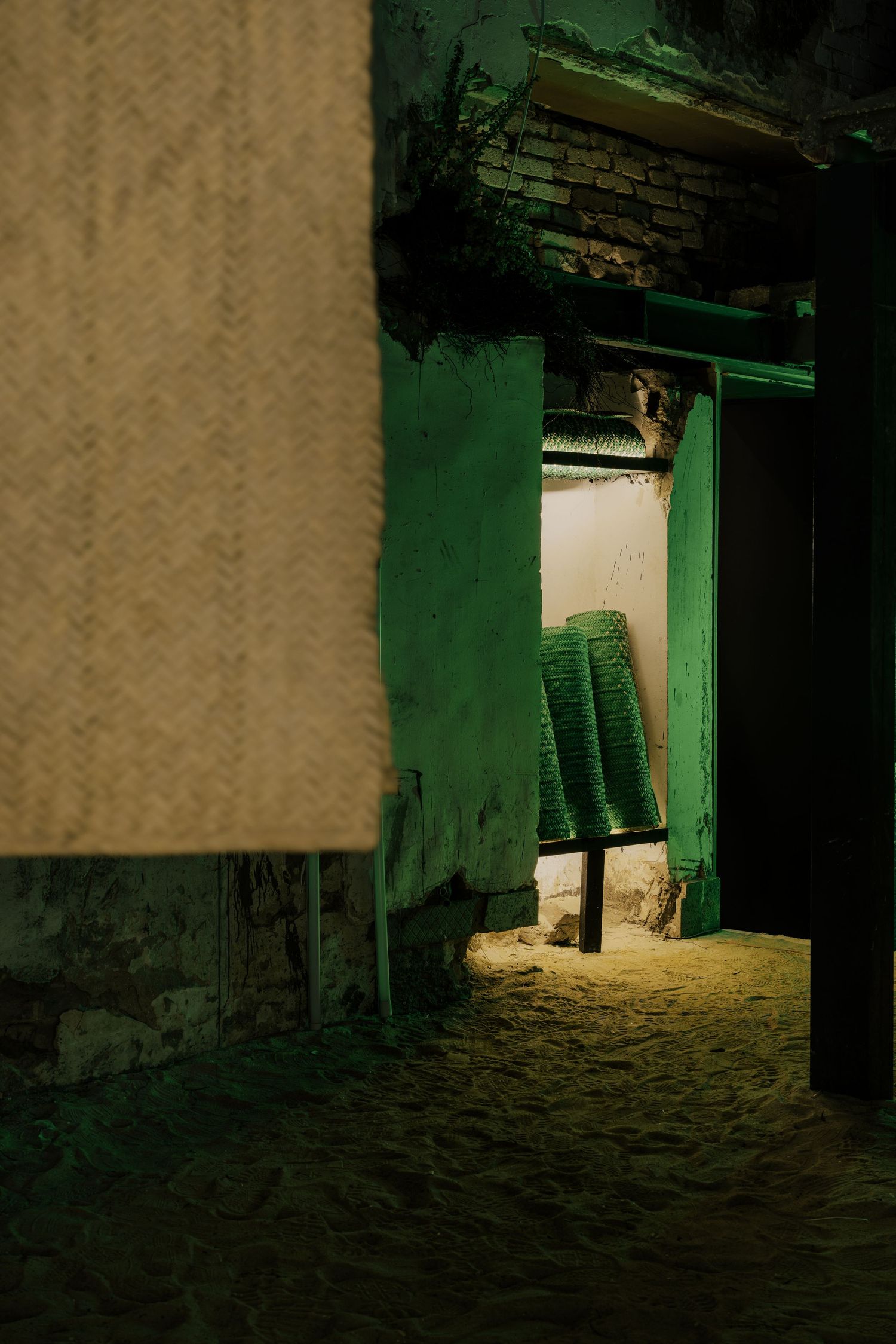 Dimly lit passageway with green walls, sandy floor, woven panel, and stacked green chairs visible.
