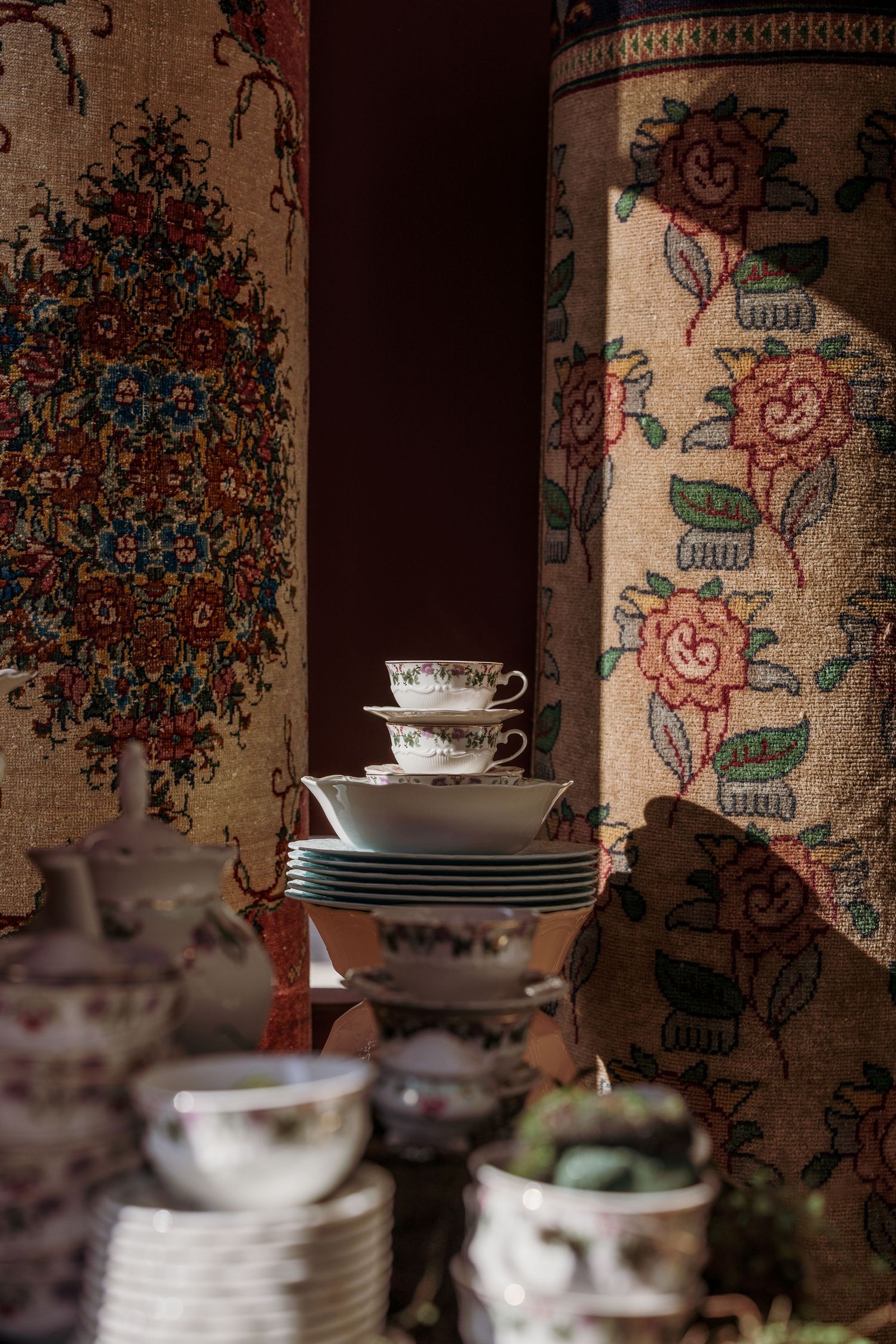 Stacked fine china teacups and saucers displayed against ornate floral tapestries in warm vintage setting.