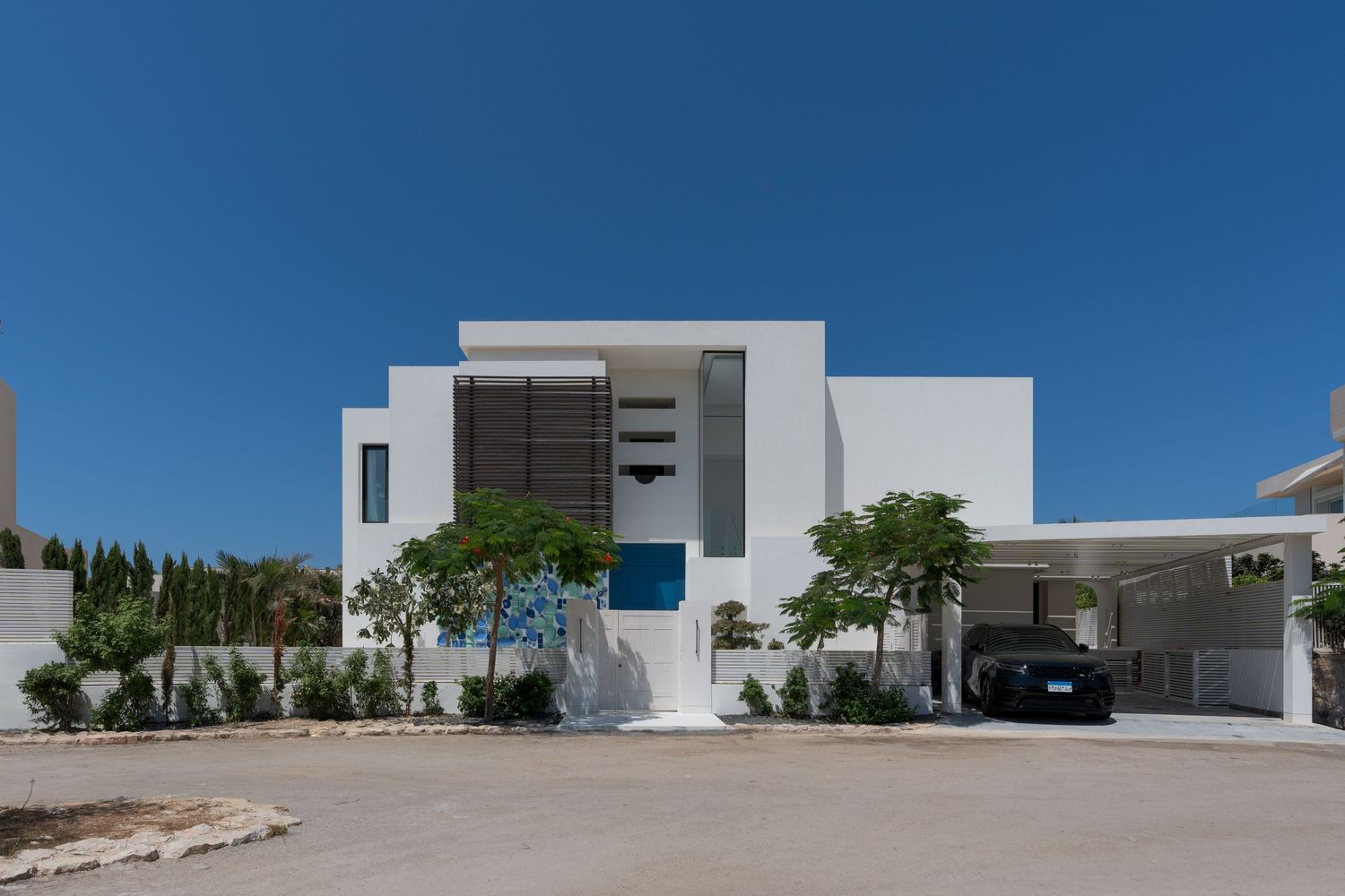Modern white two-story luxury villa with blue front door, carport, and manicured landscaping under clear blue sky.
