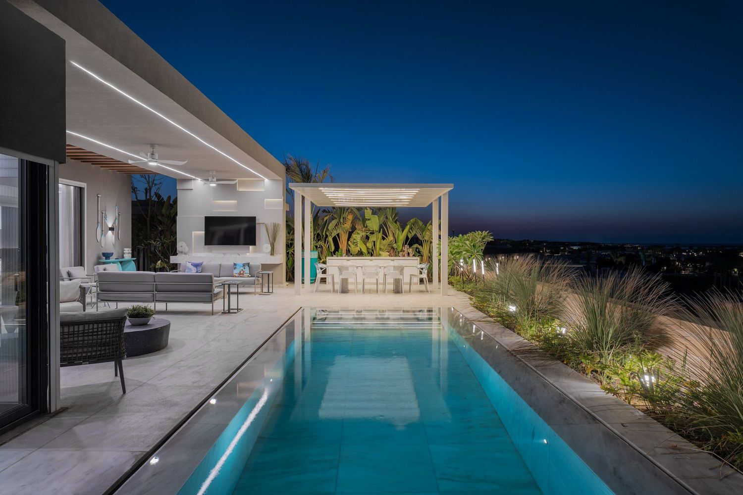 Modern villa with glowing pool, pergola, outdoor seating, and panoramic city lights at night.