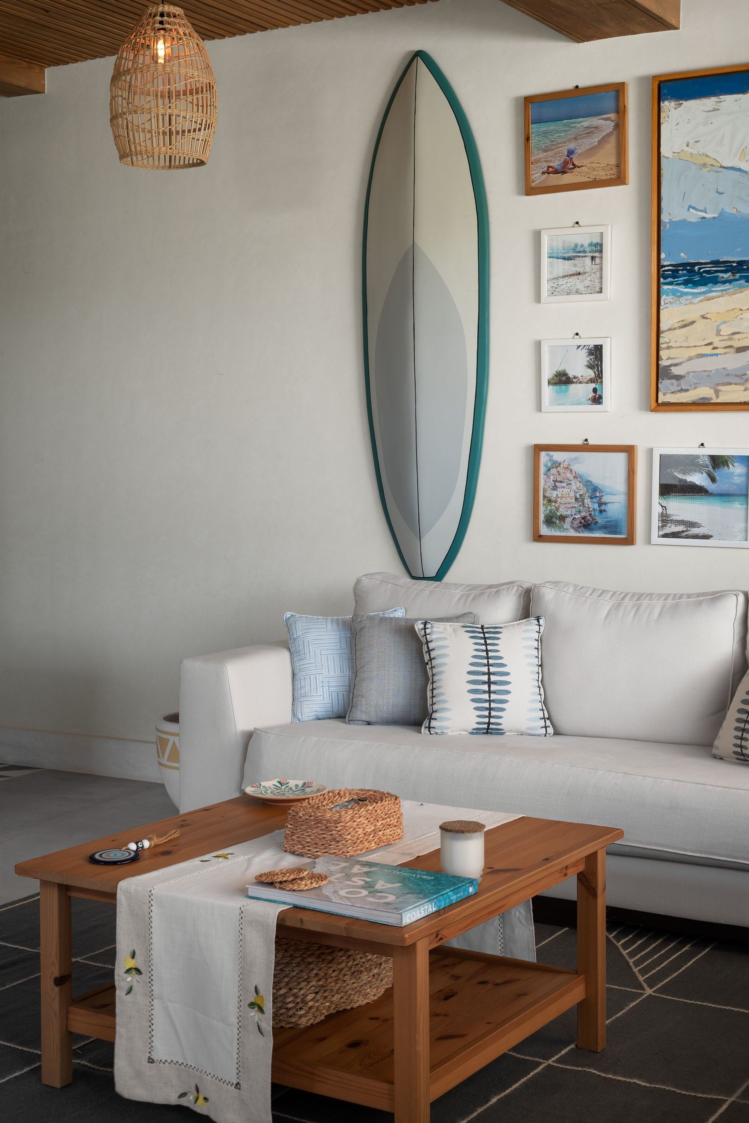 Coastal living room with surfboard mirror, beach artwork gallery wall, white sofa and wooden coffee table.