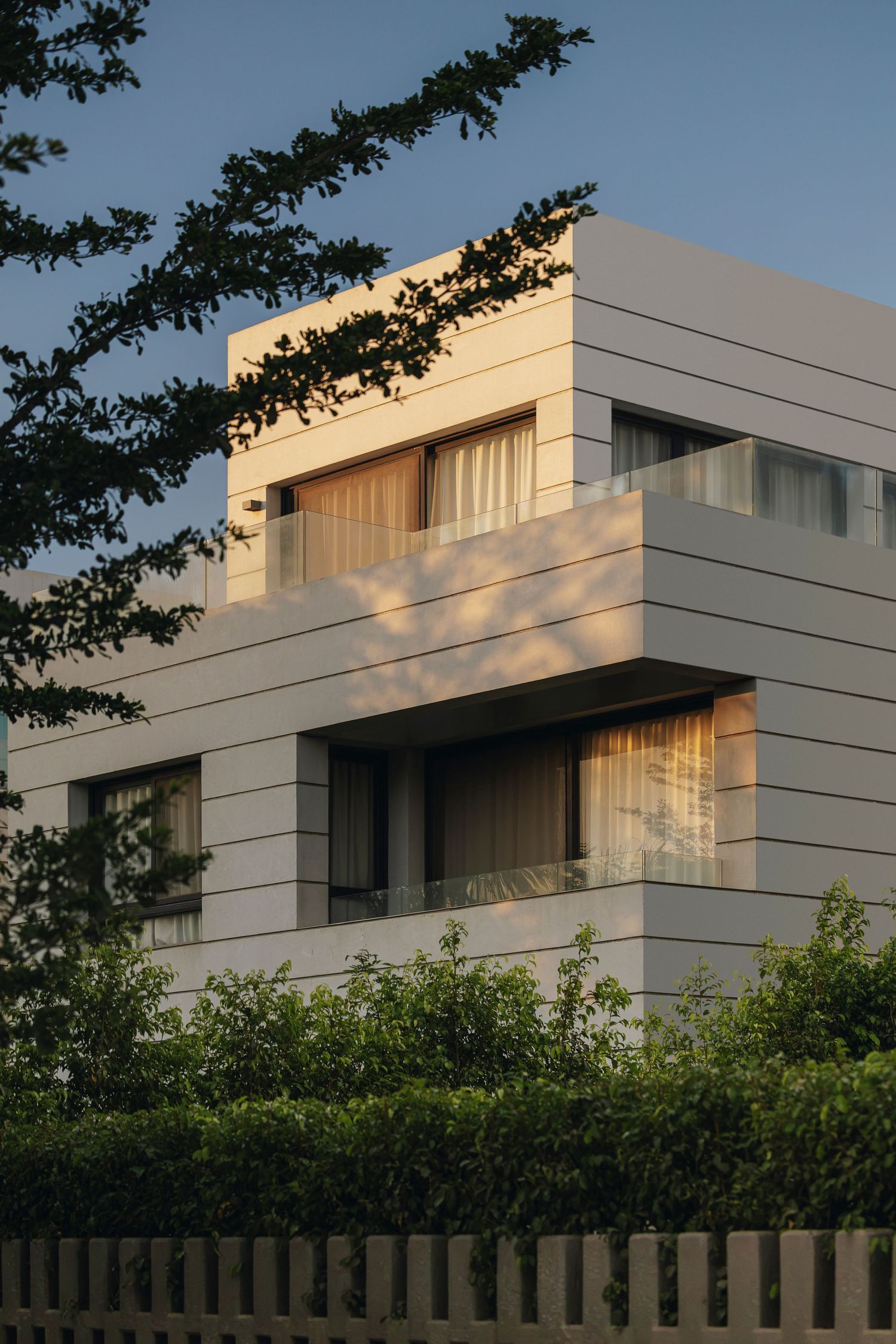 Modern white multi-story residential building with large windows, balconies, and lush green hedges at golden hour.