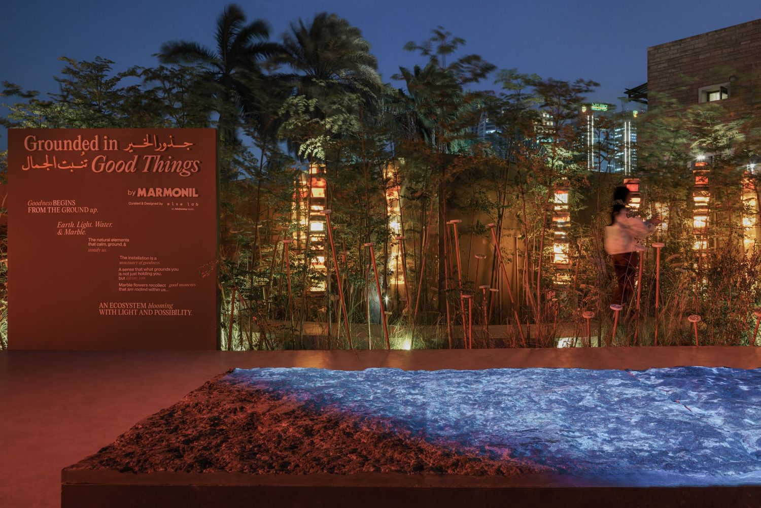 Grounded in Good Things installation by Raknomi lit up at night with red soil, blue glowing pool, and bamboo.