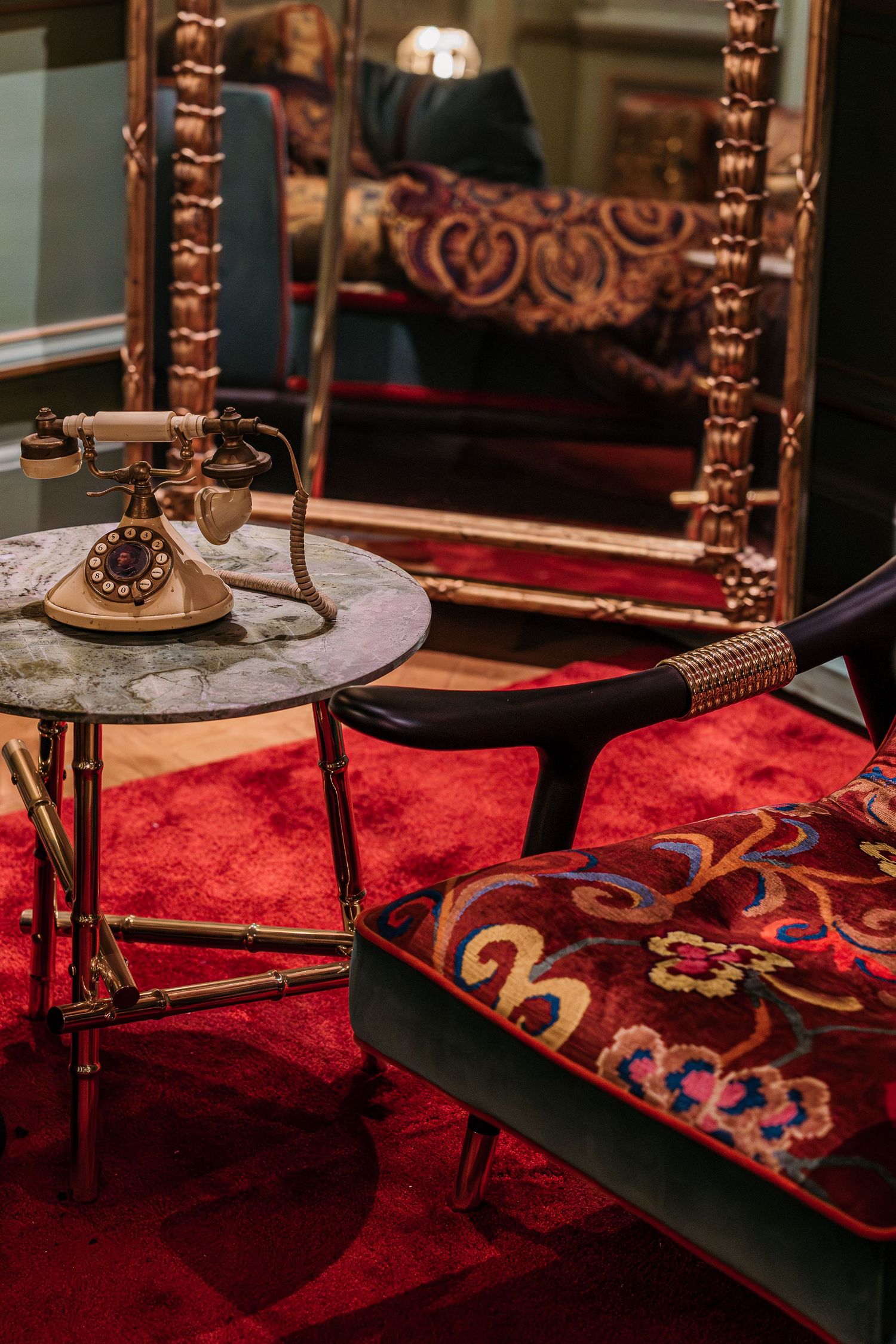 Antique rotary phone on marble brass table beside ornate mirror reflecting red velvet floral armchair interior.