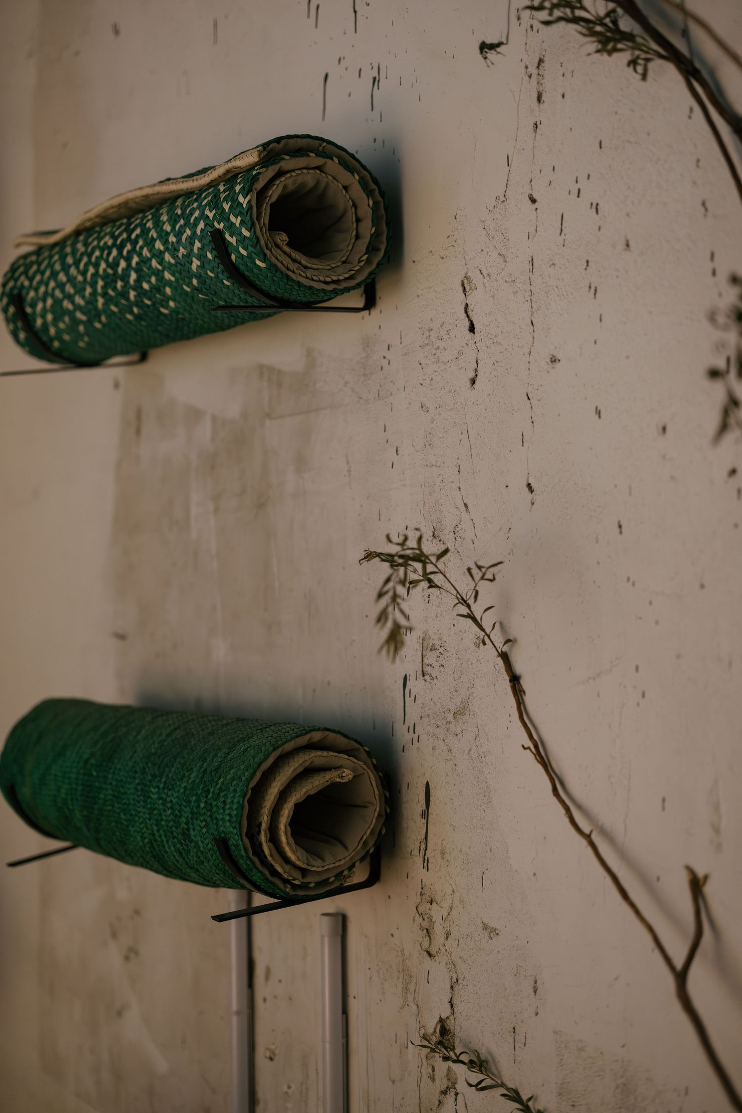 Two green rolled yoga mats mounted on wall brackets against a weathered white wall with dried plant branches.