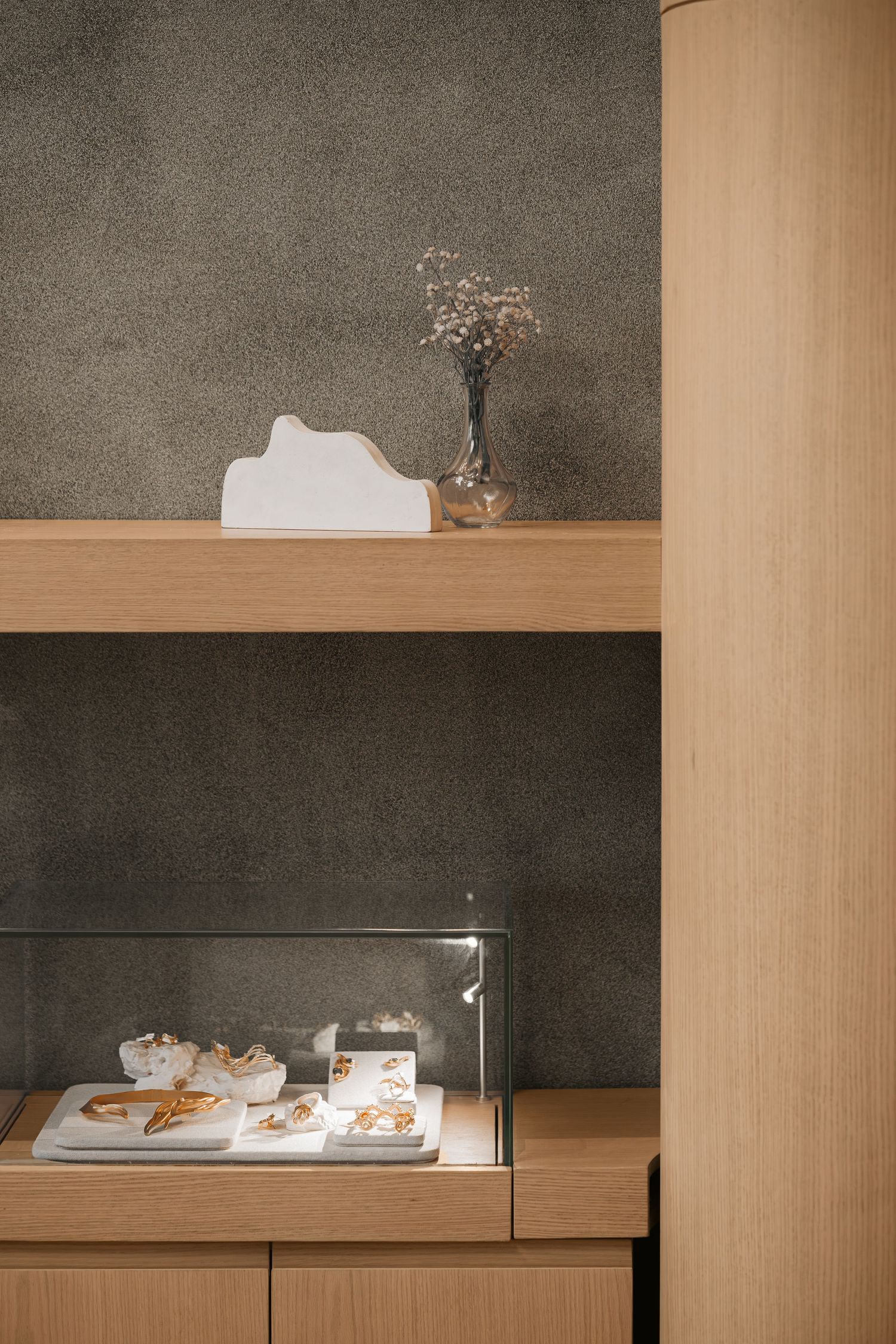 Minimalist wooden shelving display with glass case showing pastries, dried flowers, and white decorative object against gray wall.