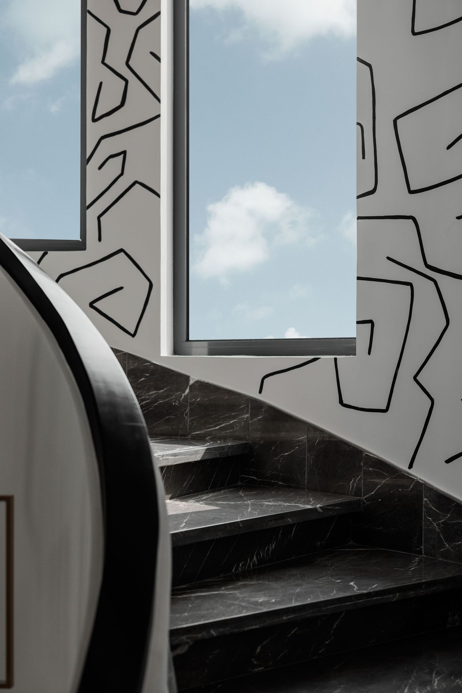 Dark marble spiral staircase with bold black line art wallpaper and large window revealing blue sky.