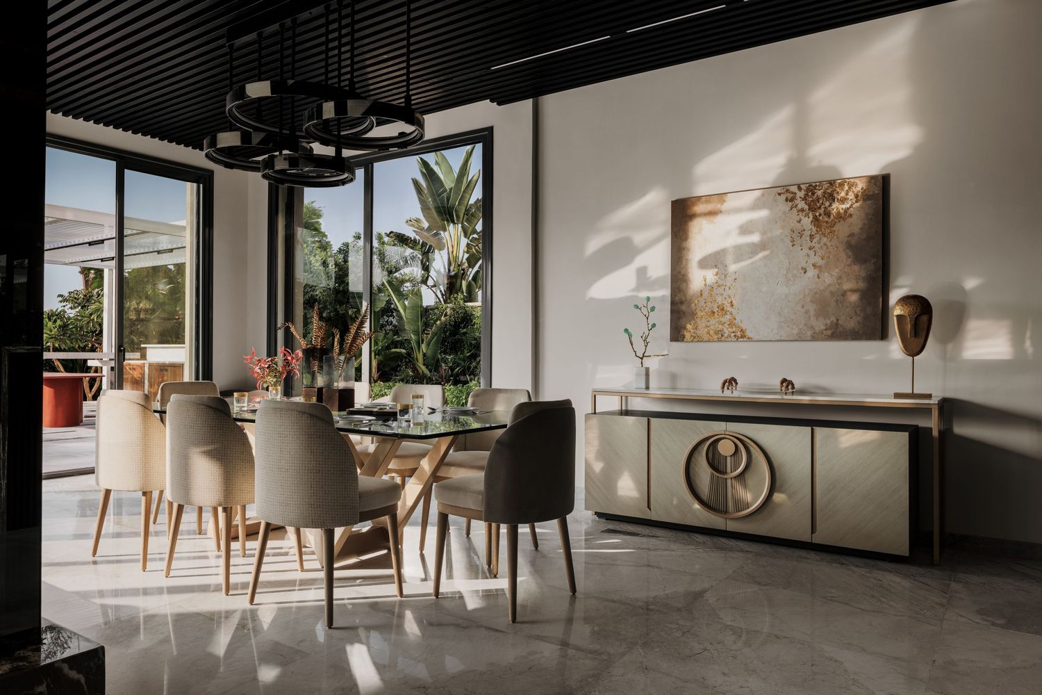 Elegant modern dining room with oval table, beige chairs, sideboard, abstract art, and floor-to-ceiling windows overlooking lush greenery.