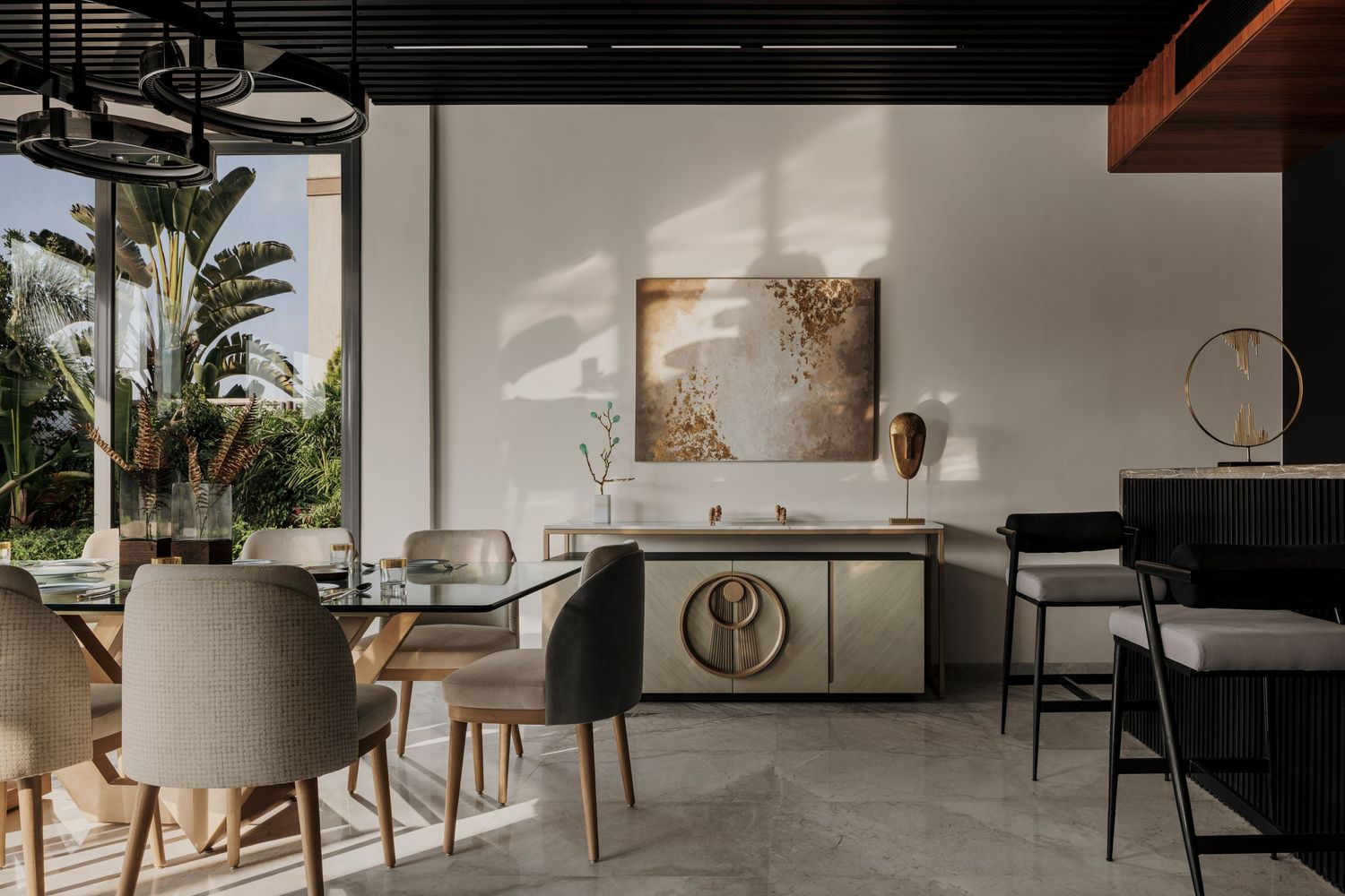 Elegant dining room with upholstered chairs, marble floors, gold cabinet, abstract artwork, and lush garden views.