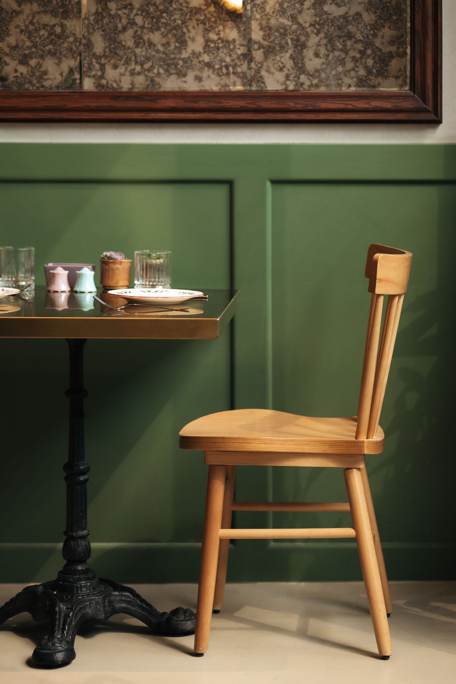 Wooden bistro chair beside a brass-top table against a green paneled wall with ornate mirror above.