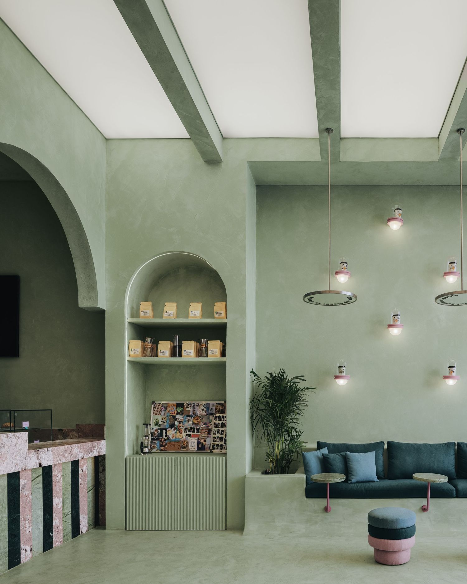 Sage green cafe interior with arched alcove shelving, terrazzo counter, blue sofa, and decorative pendant lights.