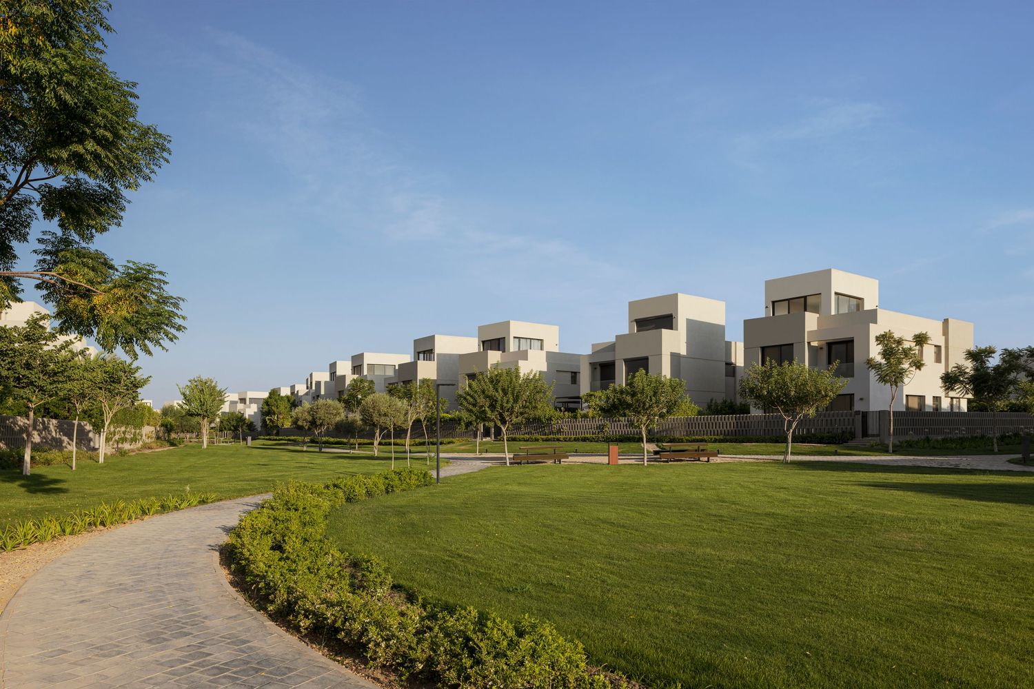 Modern white cubic villas surrounded by lush green lawns and manicured hedges under a clear blue sky.