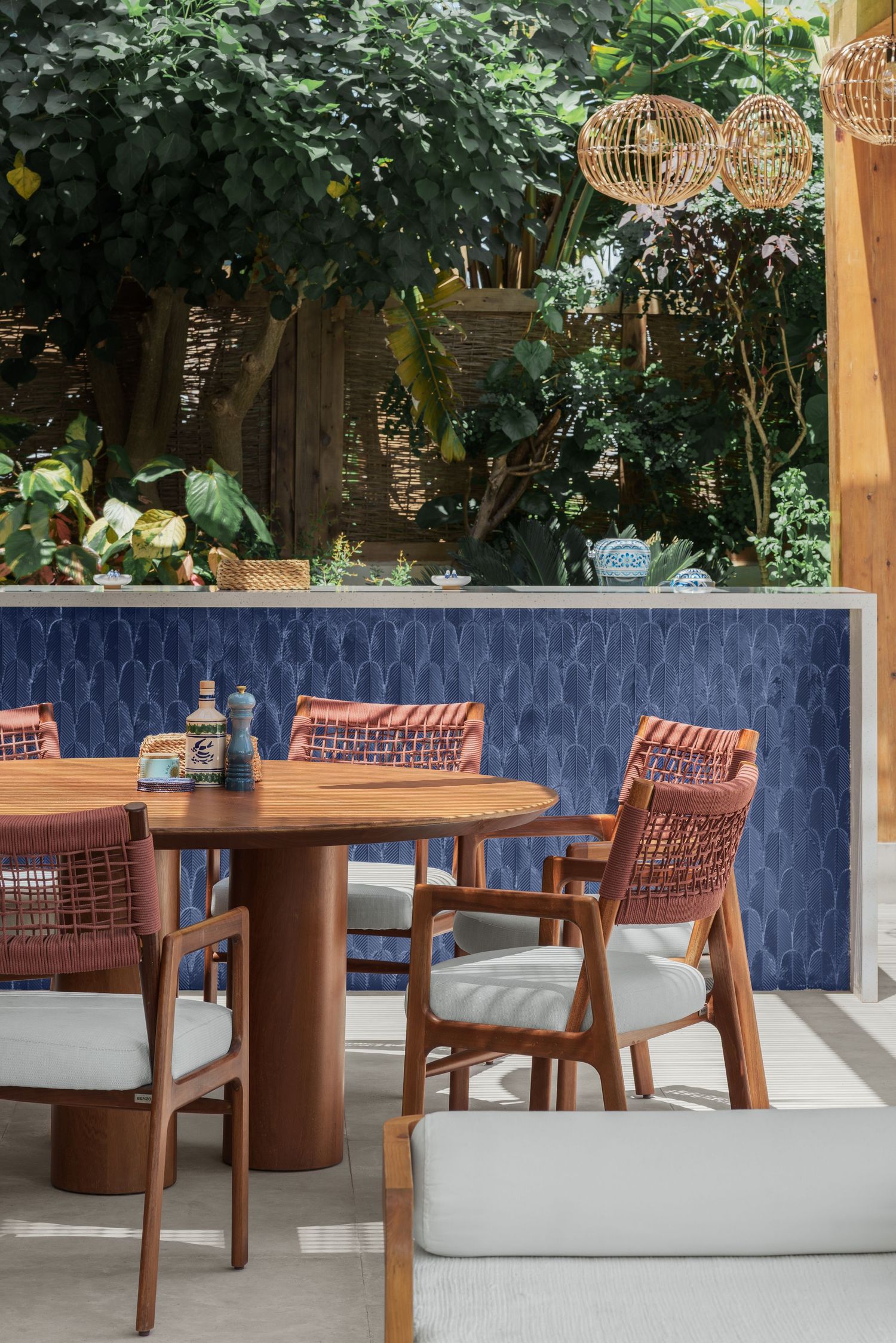 Al fresco dining setup with round wooden table, woven chairs, blue mosaic wall, and tropical garden.