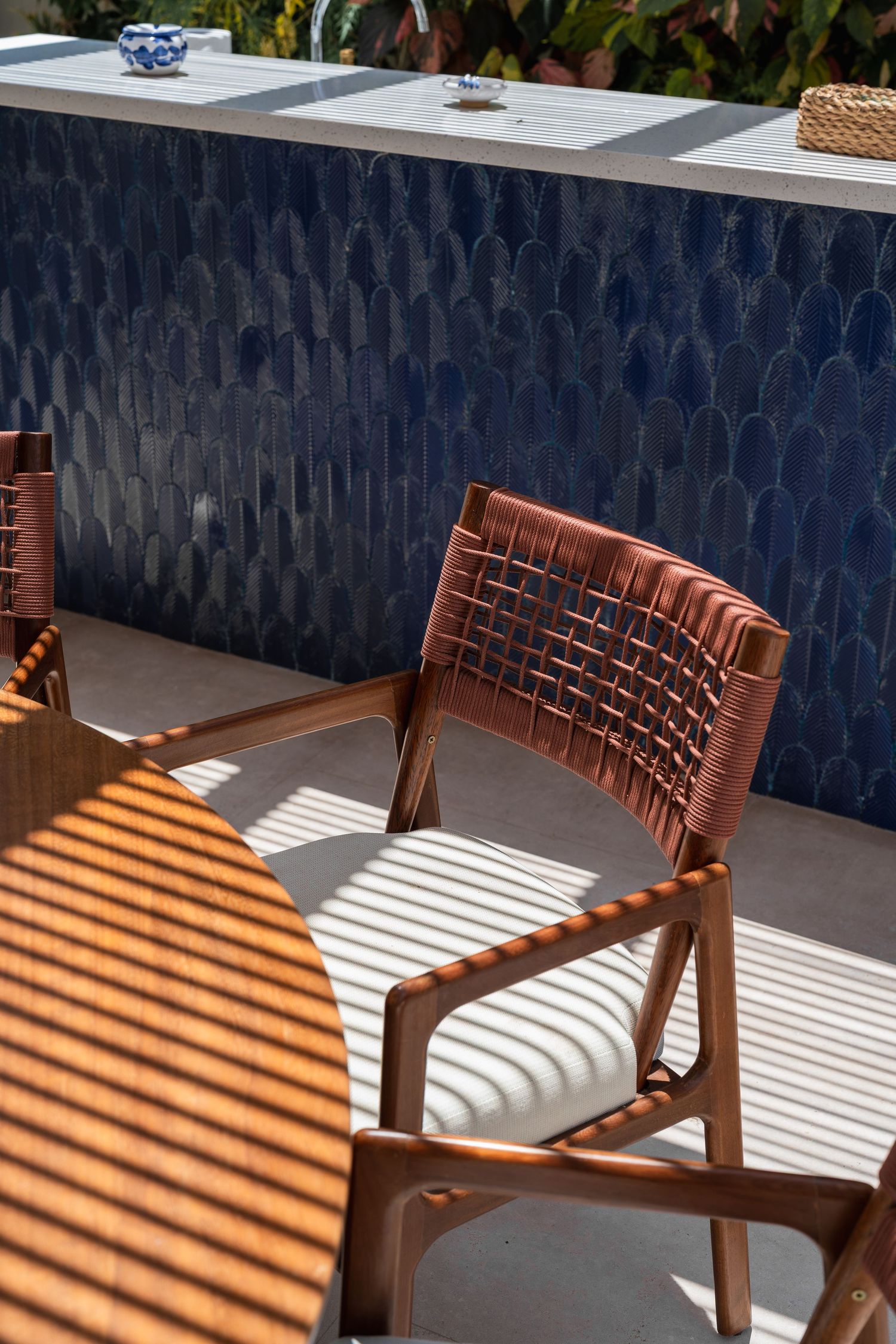 Outdoor dining chair with woven back beside blue mosaic tiled wall in sunlit patio setting.