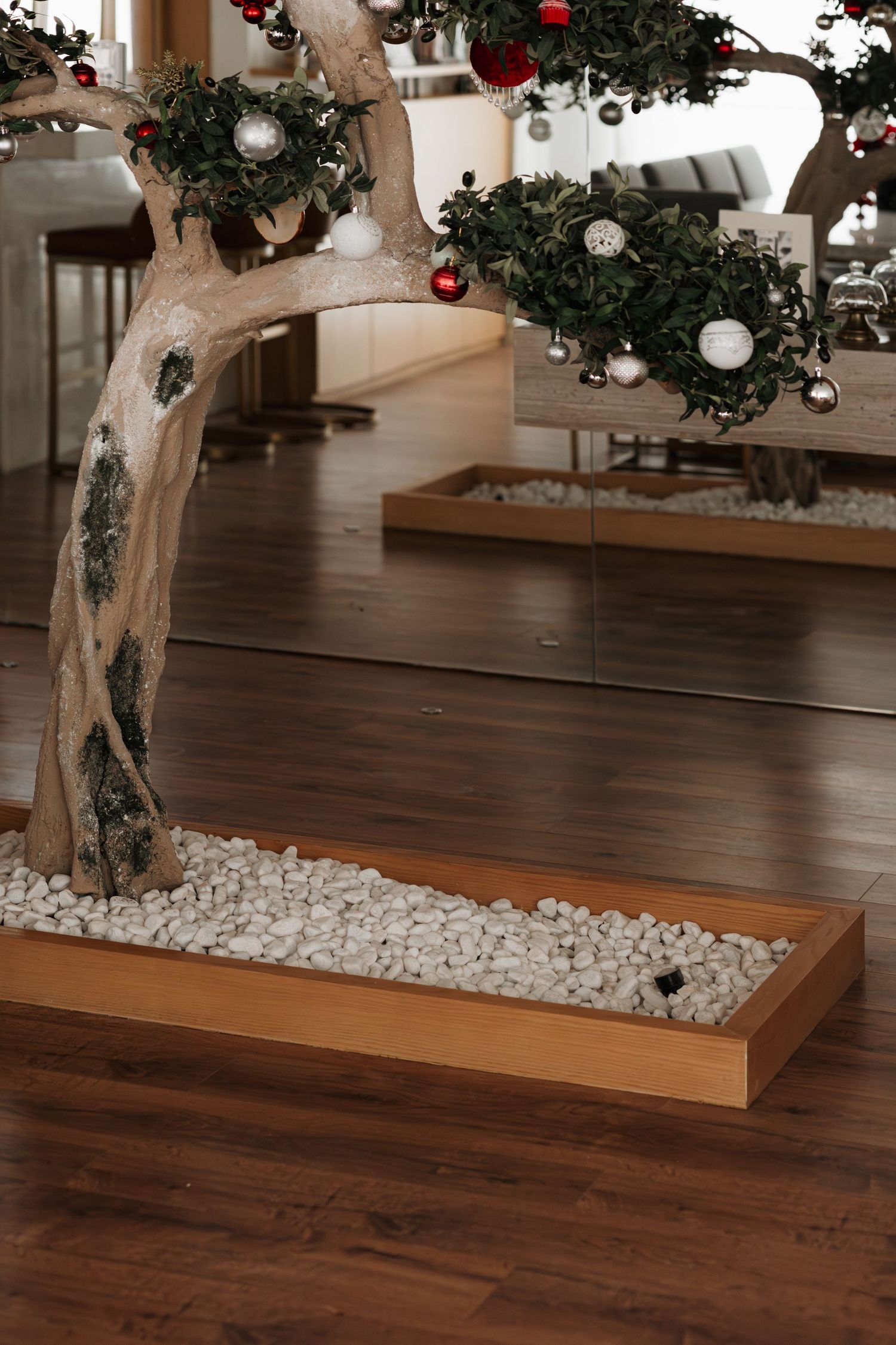 Decorative indoor bonsai-style tree with Christmas ornaments in white pebble planter box on dark hardwood floor.