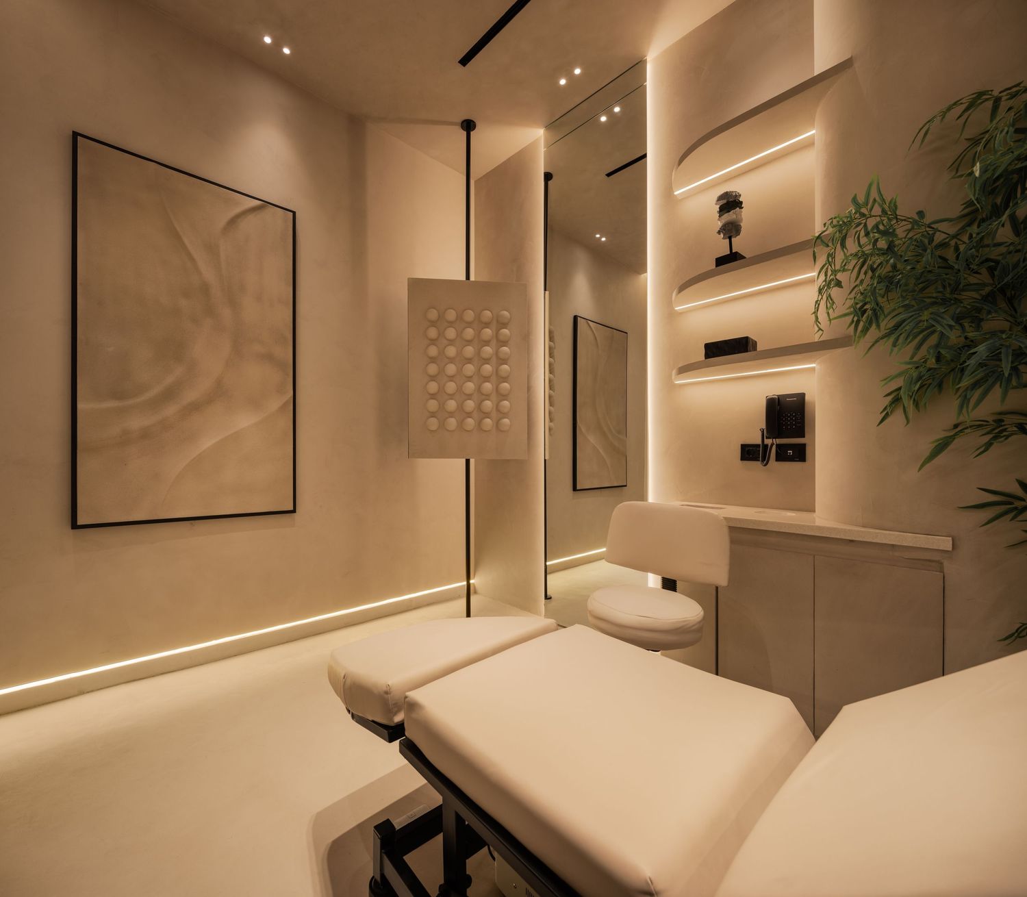Minimalist luxury treatment room with white massage bed, warm LED lighting, built-in shelves, and bamboo plant.