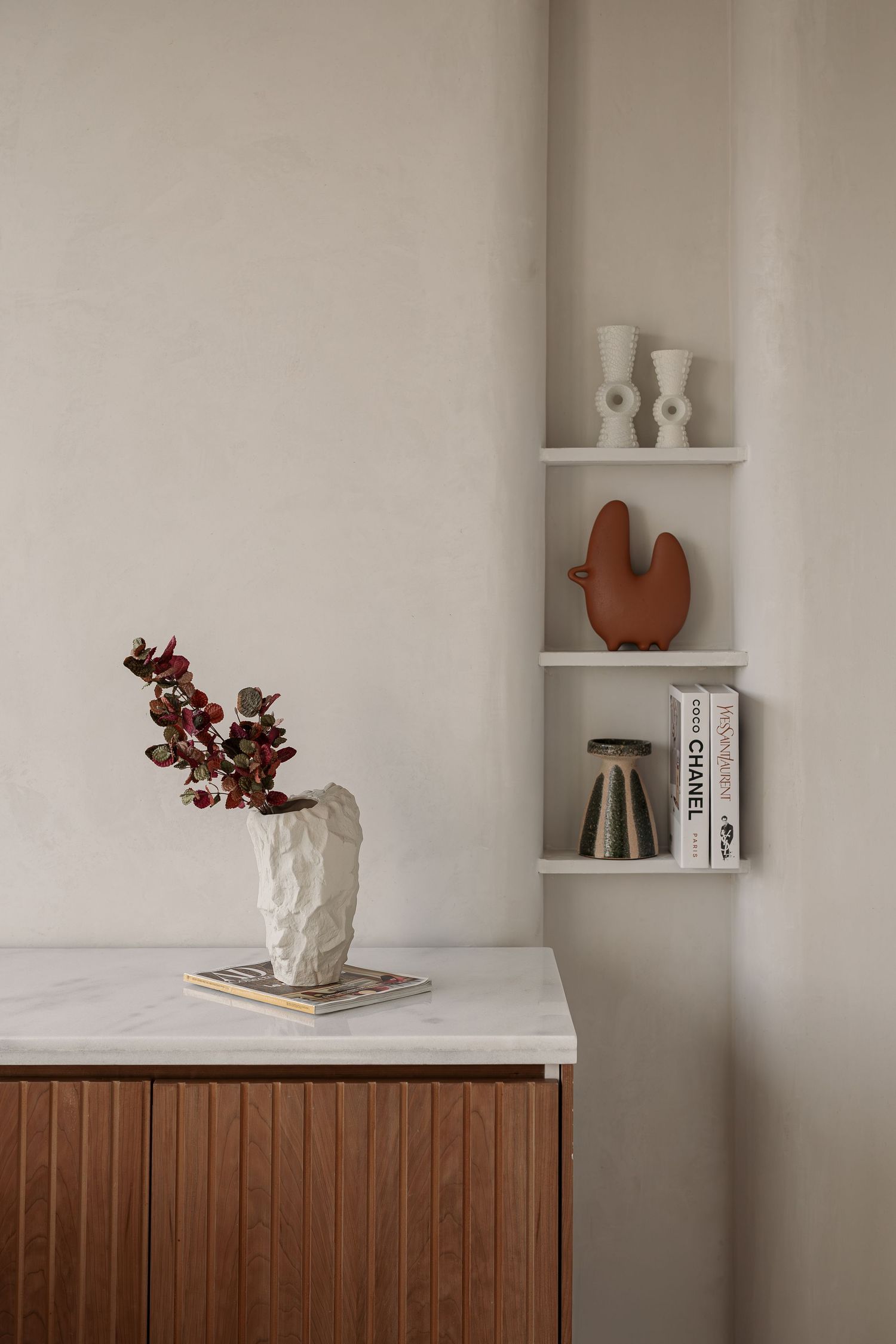 Minimalist interior with wooden cabinet, marble top, textured vase with flowers, and corner wall shelves.