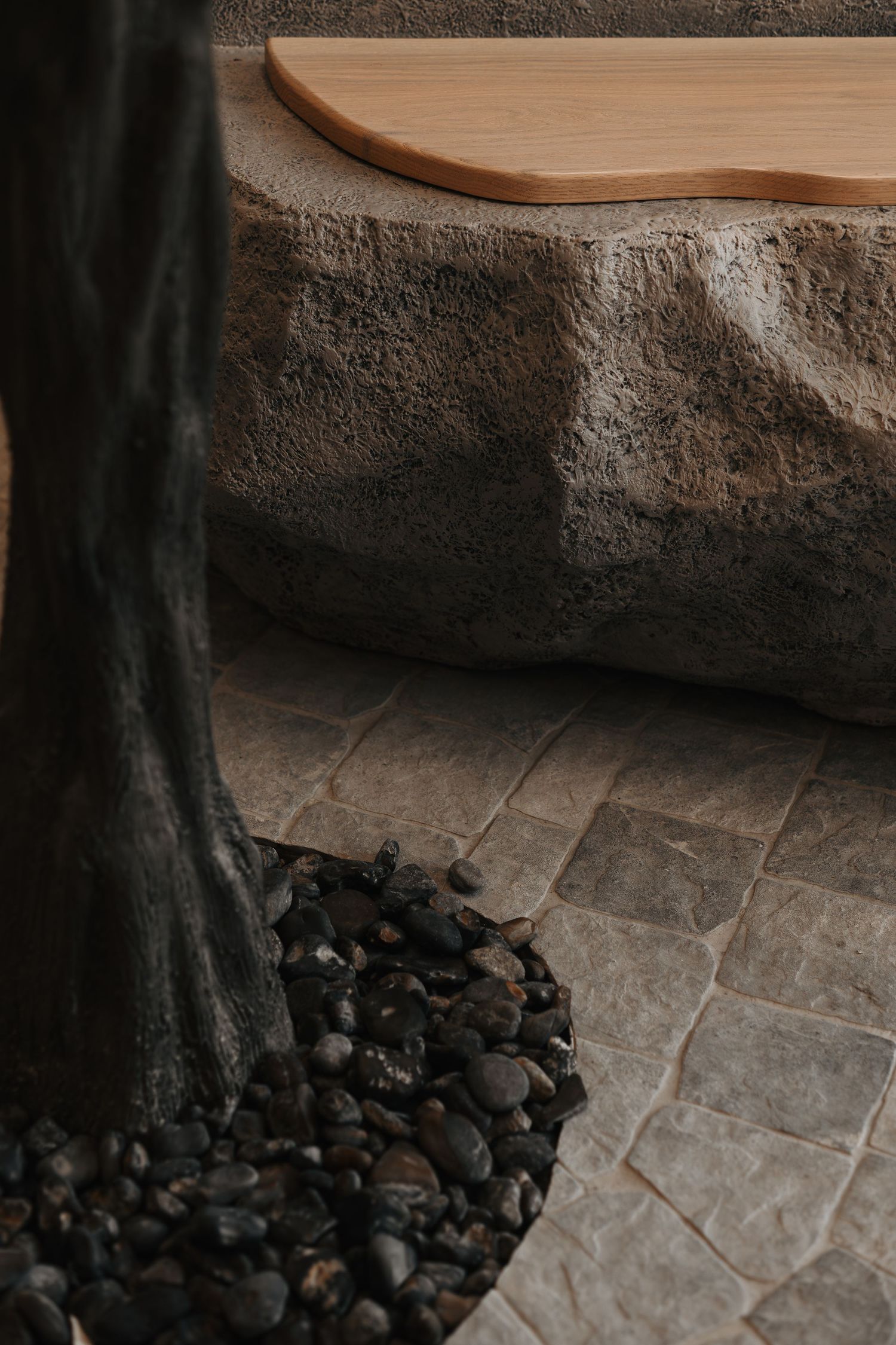 Natural stone bench with wooden top surrounded by pebbles and olive tree trunk on textured tile flooring.