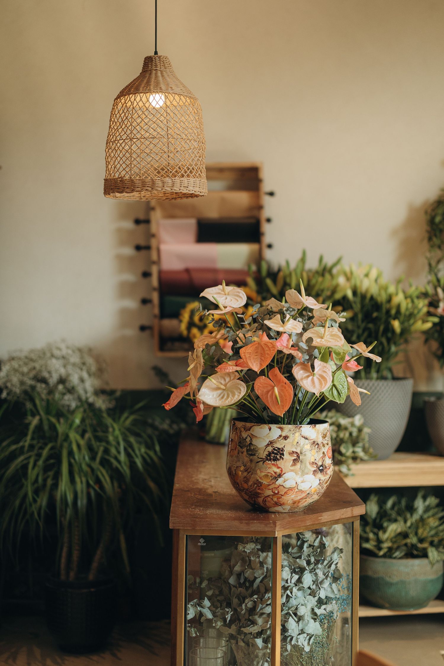 Boho-style interior with wicker pendant light, orange anthurium flowers in decorative pot, and lush green plants on wooden table.