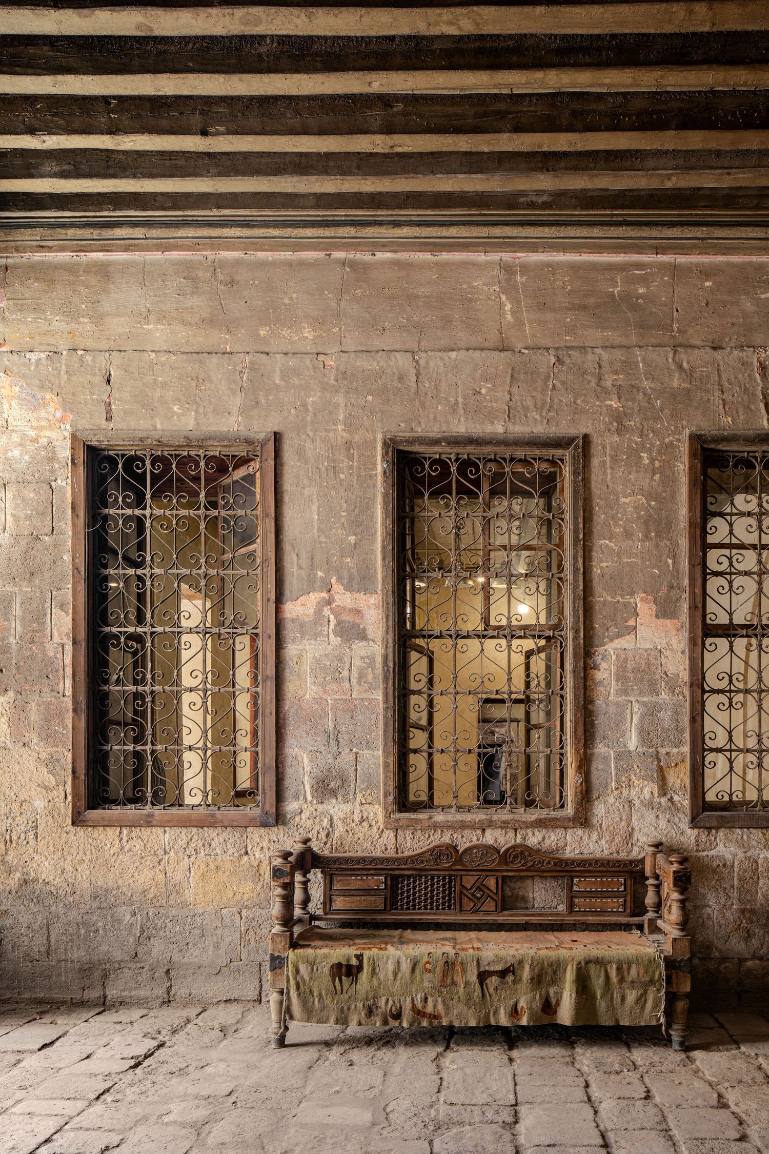 Ancient stone corridor with ornate iron-grilled windows, weathered walls, and an antique carved wooden bench.