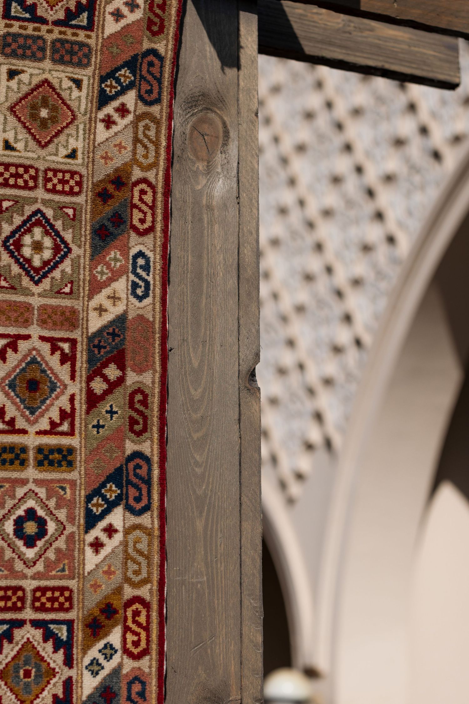 Traditional geometric patterned carpet beside weathered wooden beam with arched plasterwork ceiling beyond.