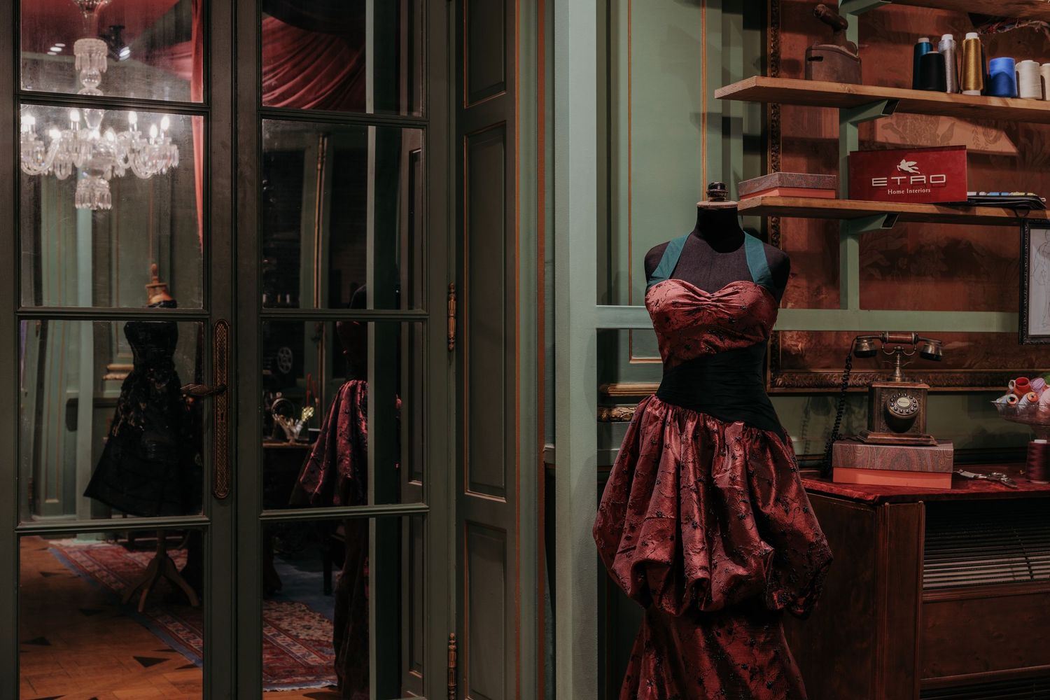 Elegant red brocade gown on mannequin in dimly lit vintage atelier with chandelier reflection in glass.