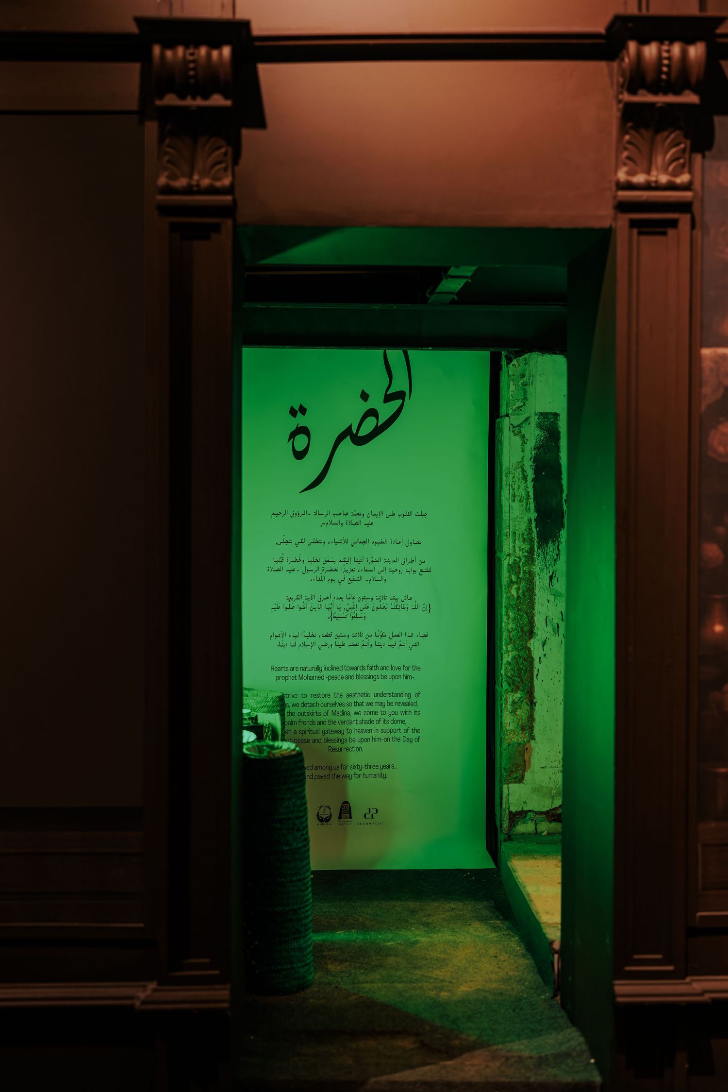 Green-lit room entrance with Arabic calligraphy text on wall, viewed through ornate dark doorframe.
