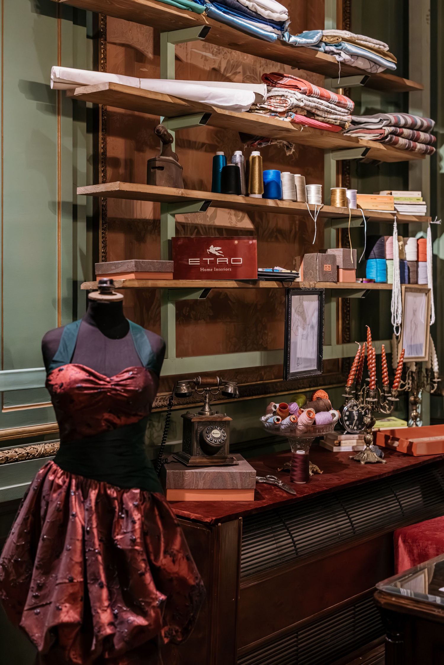 A burgundy dress on a mannequin in a fashion atelier with fabric rolls and sewing supplies on wooden shelves.