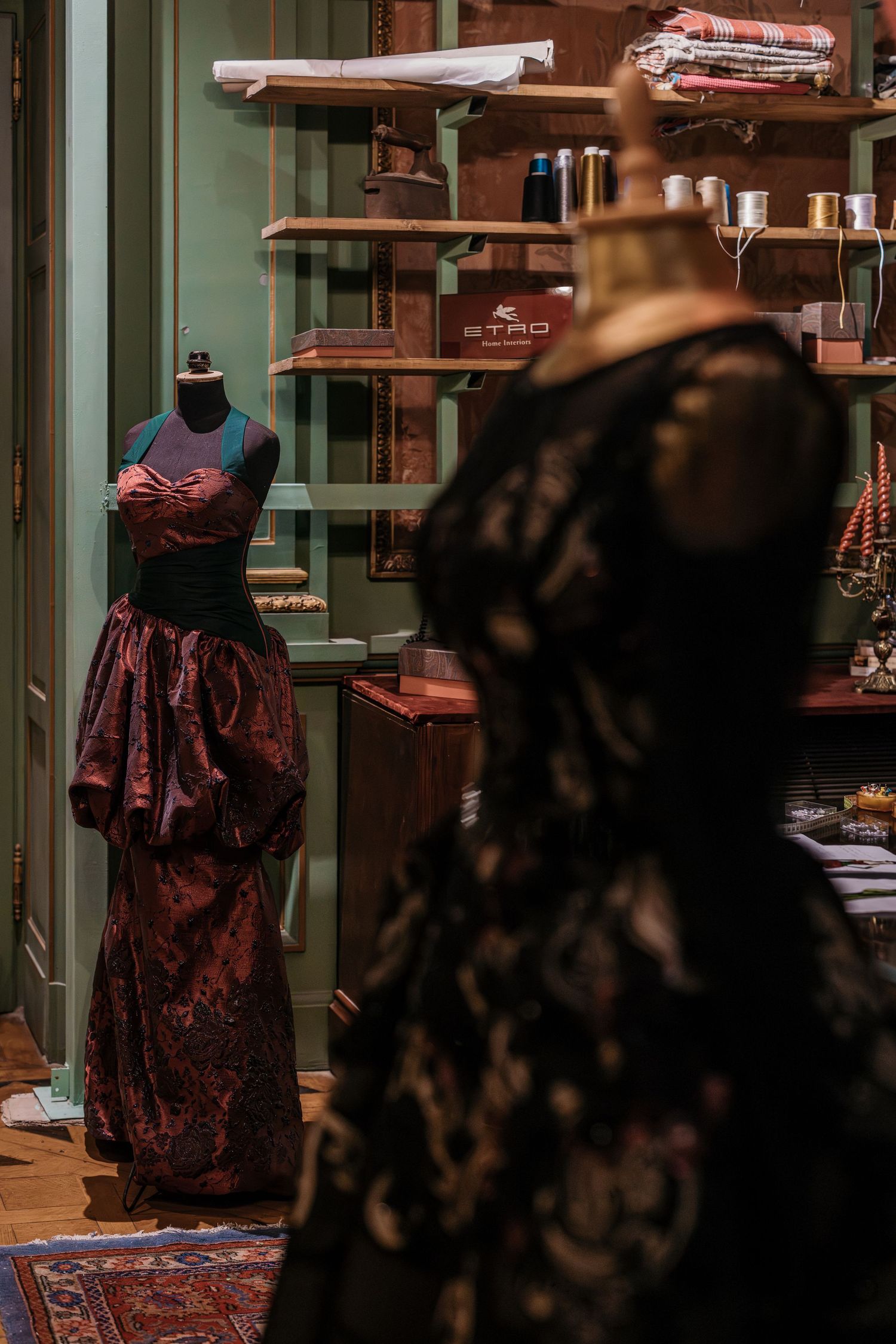 Two mannequins display elegant gowns in a vintage fashion atelier with wooden shelves and sewing supplies.