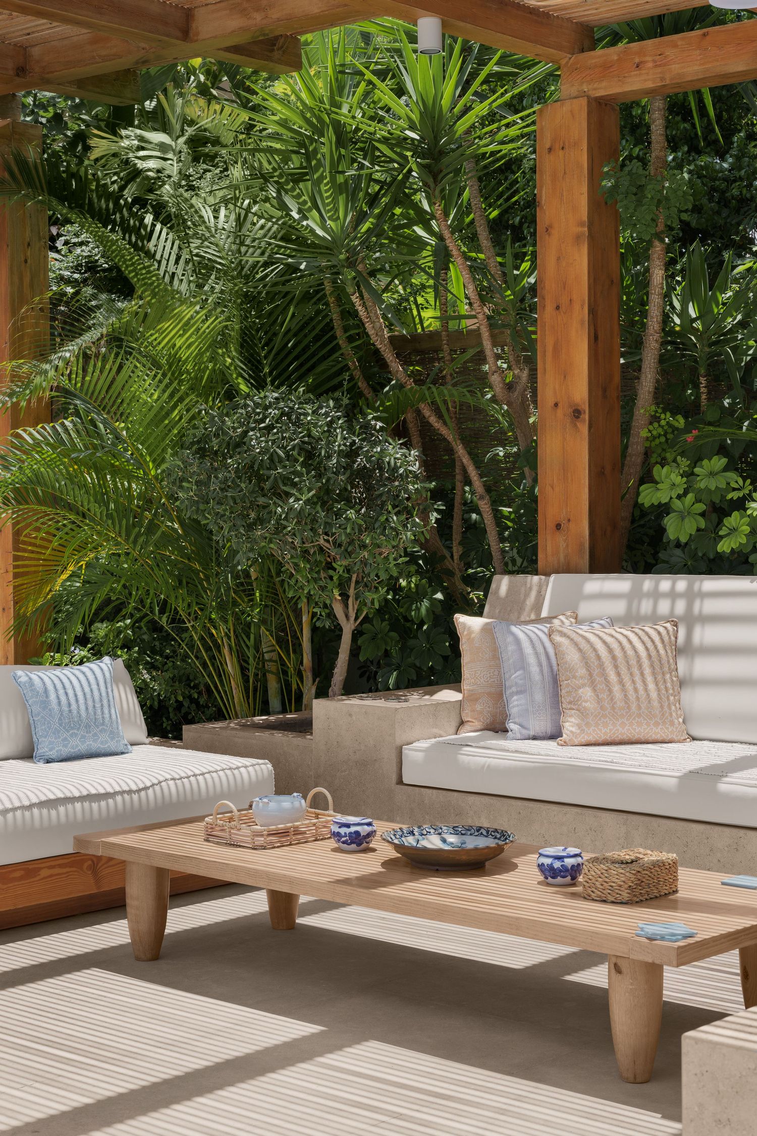 Tropical outdoor patio with wooden pergola, neutral cushioned sofas, and lush green garden backdrop.