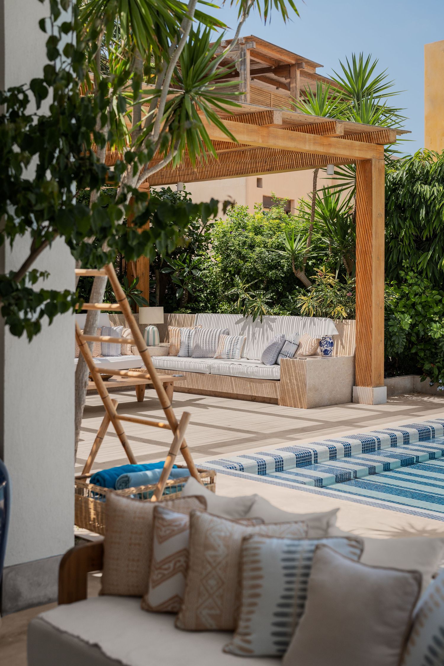 Outdoor lounge area with wooden pergola, cushioned sofa, tropical palms, and striped pool surround.