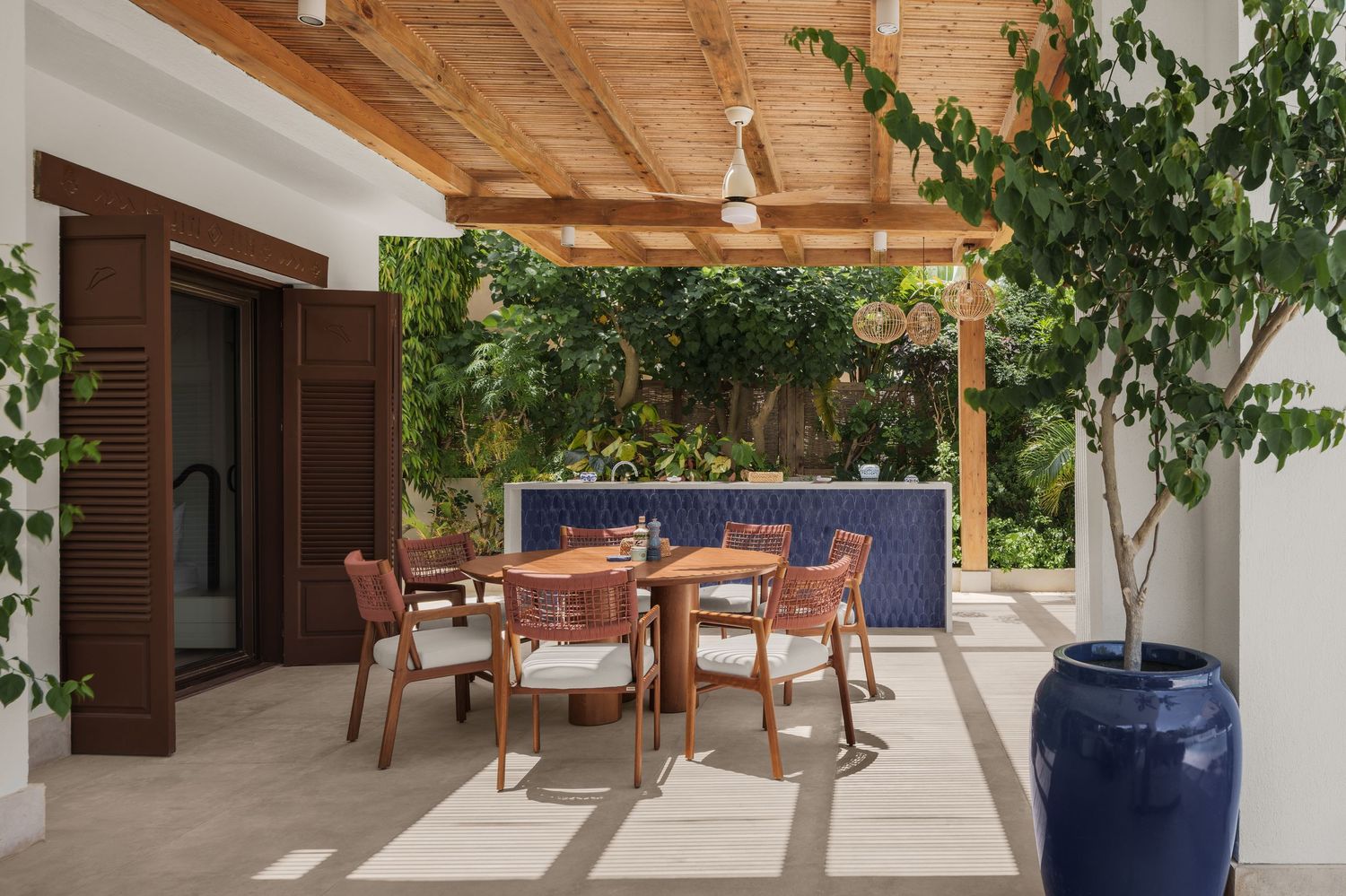 Shaded outdoor dining area with wooden pergola, rattan chairs, ceiling fan, and blue tiled accent wall.