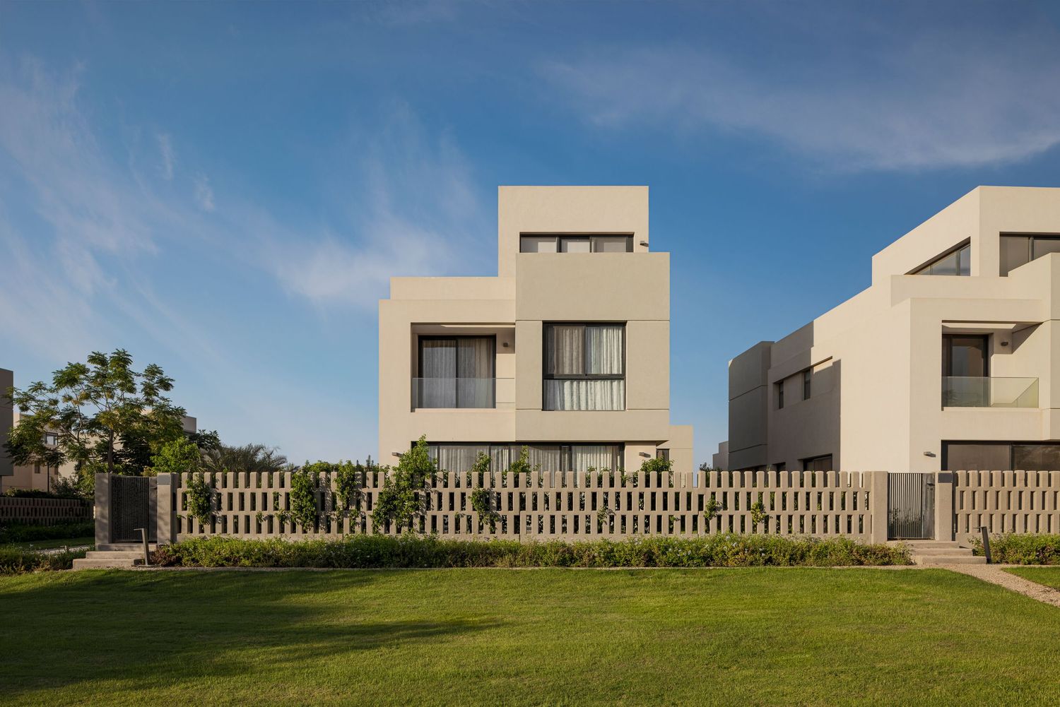 Modern white cubic villas with manicured lawn and decorative fence under blue sky.