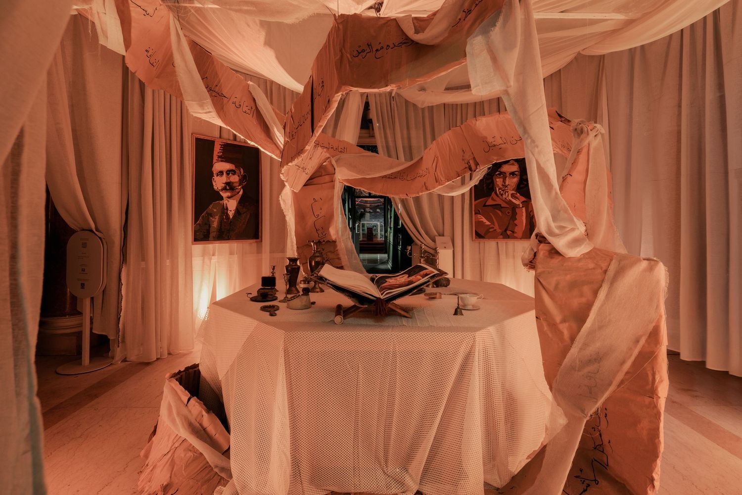 Artistic room installation with draped white and peach fabric, portraits, open books, and candles on table.