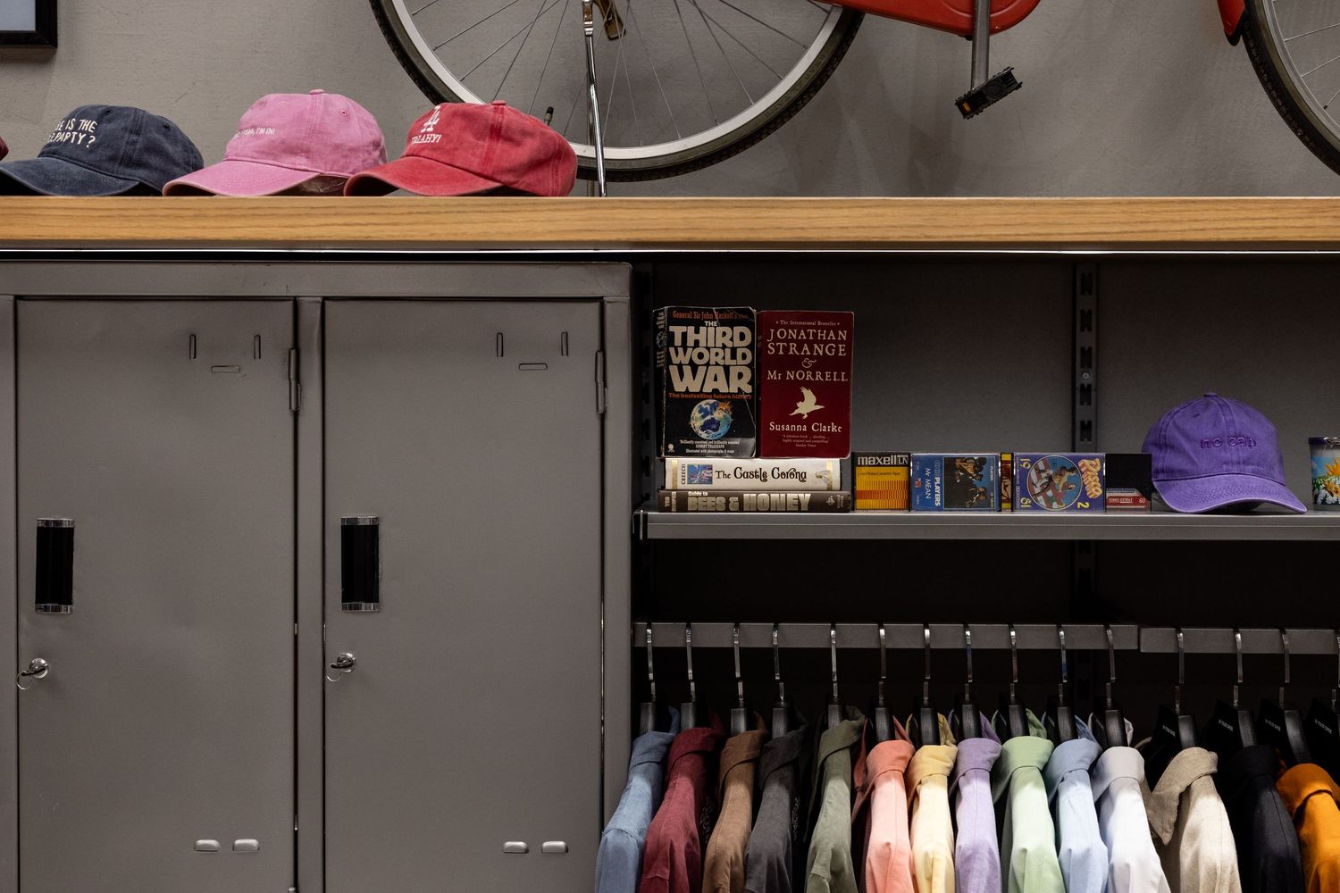 Gray metal lockers with colorful hats on top beside a clothing rack and book shelf in a boutique.