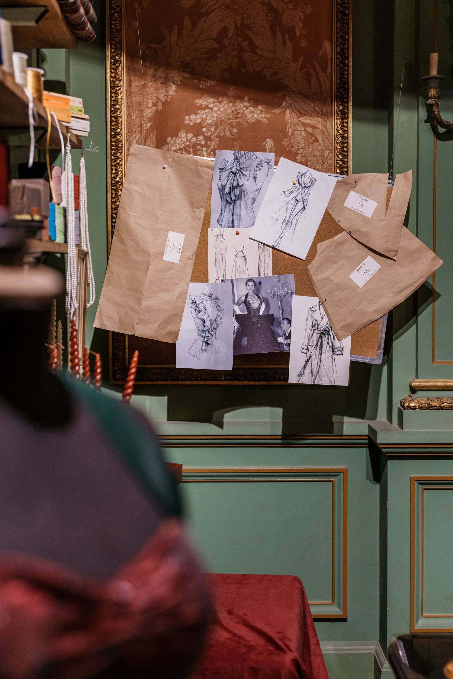 Fashion design sketches pinned to kraft paper on a green ornate wall in a couture atelier setting.