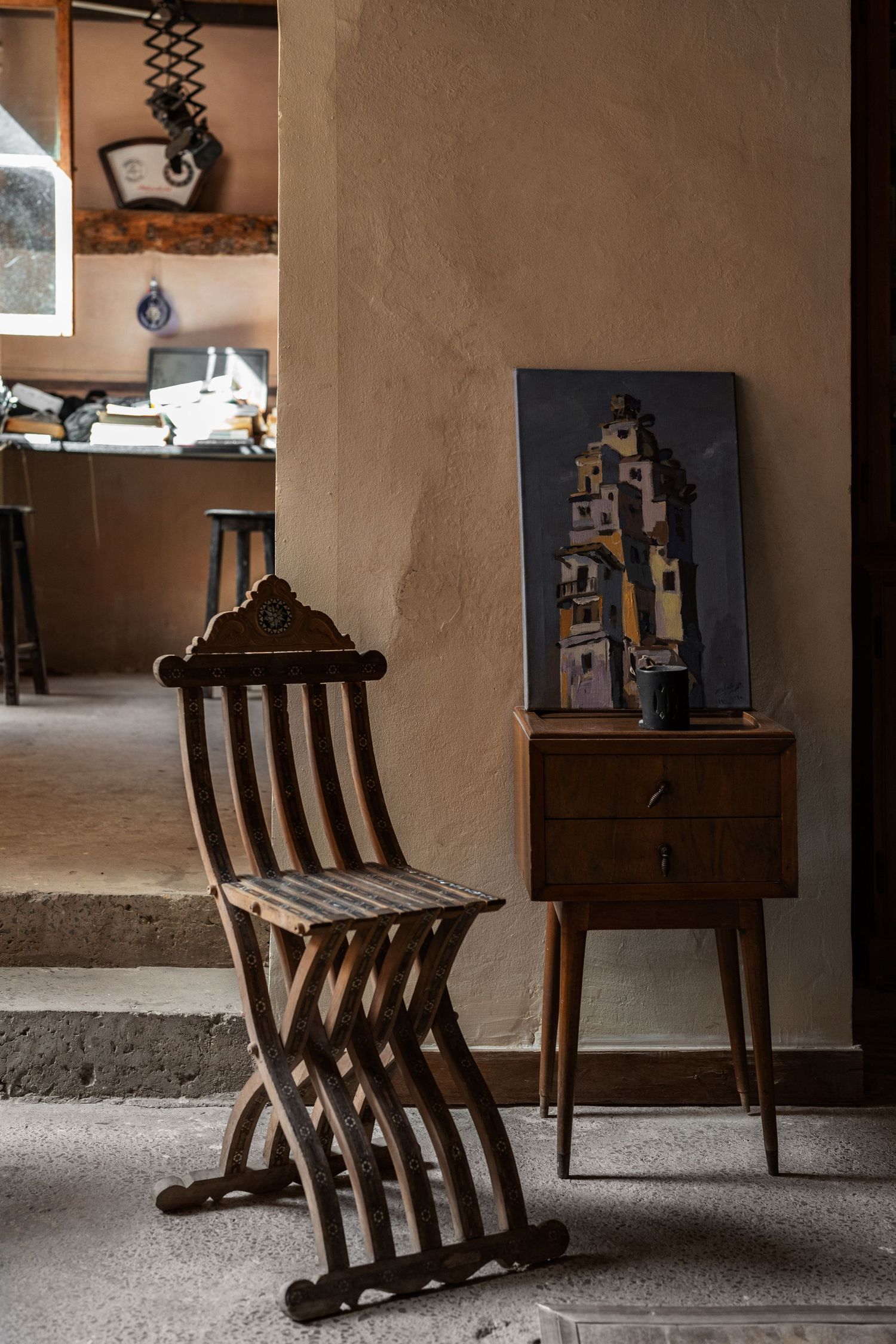 Antique wooden folding chair beside a small cabinet with a painted cityscape artwork on stone floor.