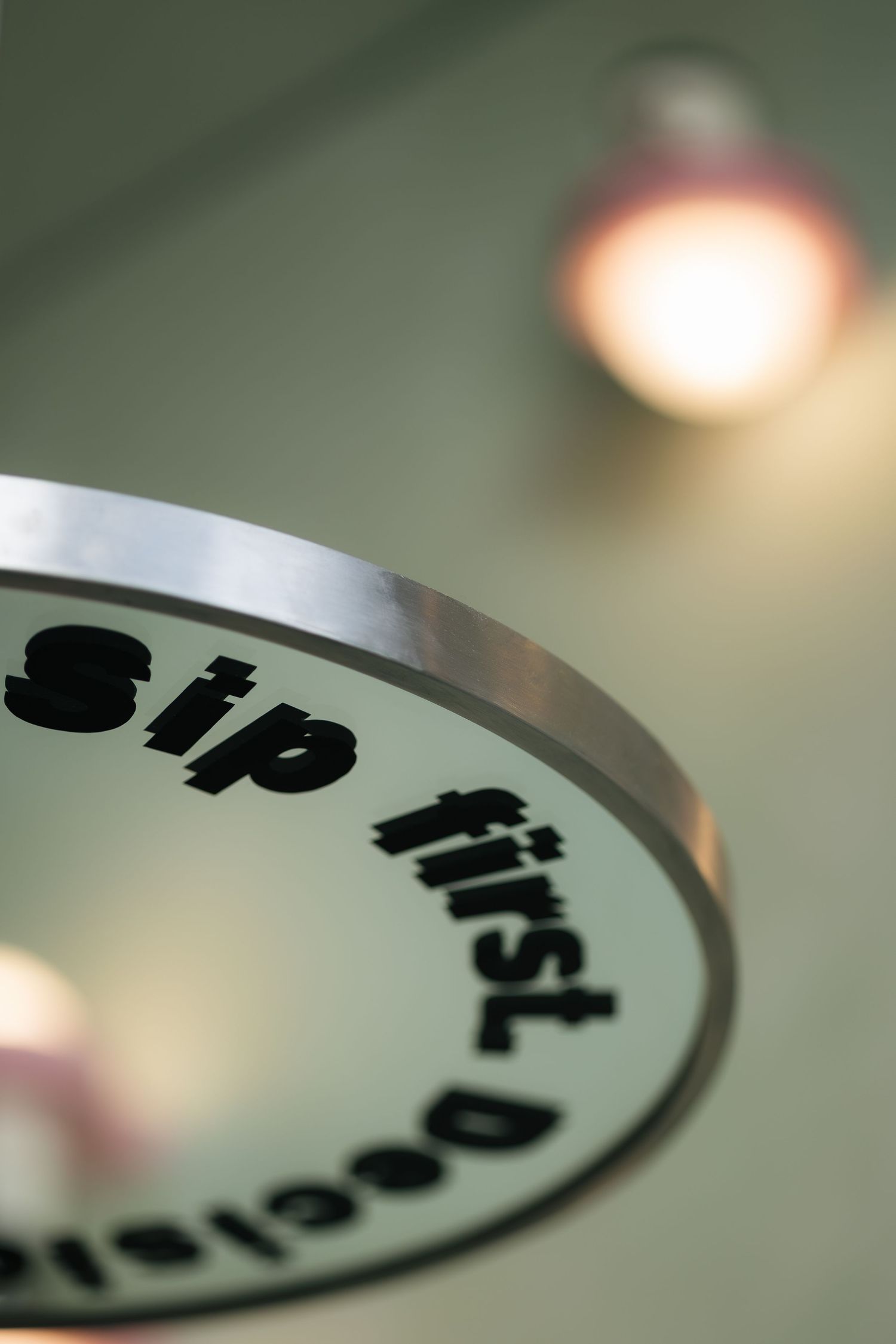 Close-up of a circular sign reading sip first decide later with blurred warm pendant lights in background.
