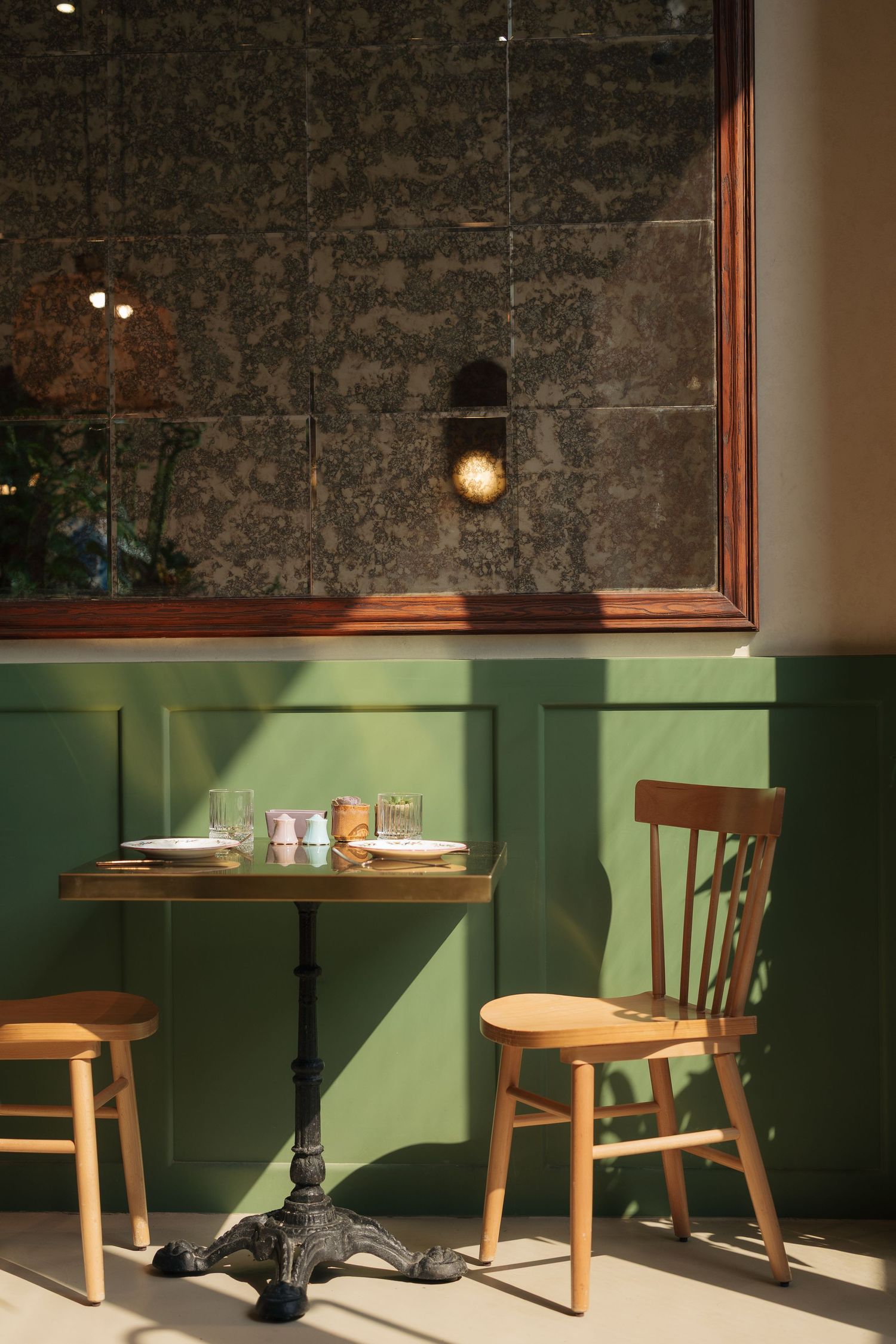 Sunlit cafe corner with green paneled wall, bistro table, wooden chairs, and large antique mirror reflecting warm light.