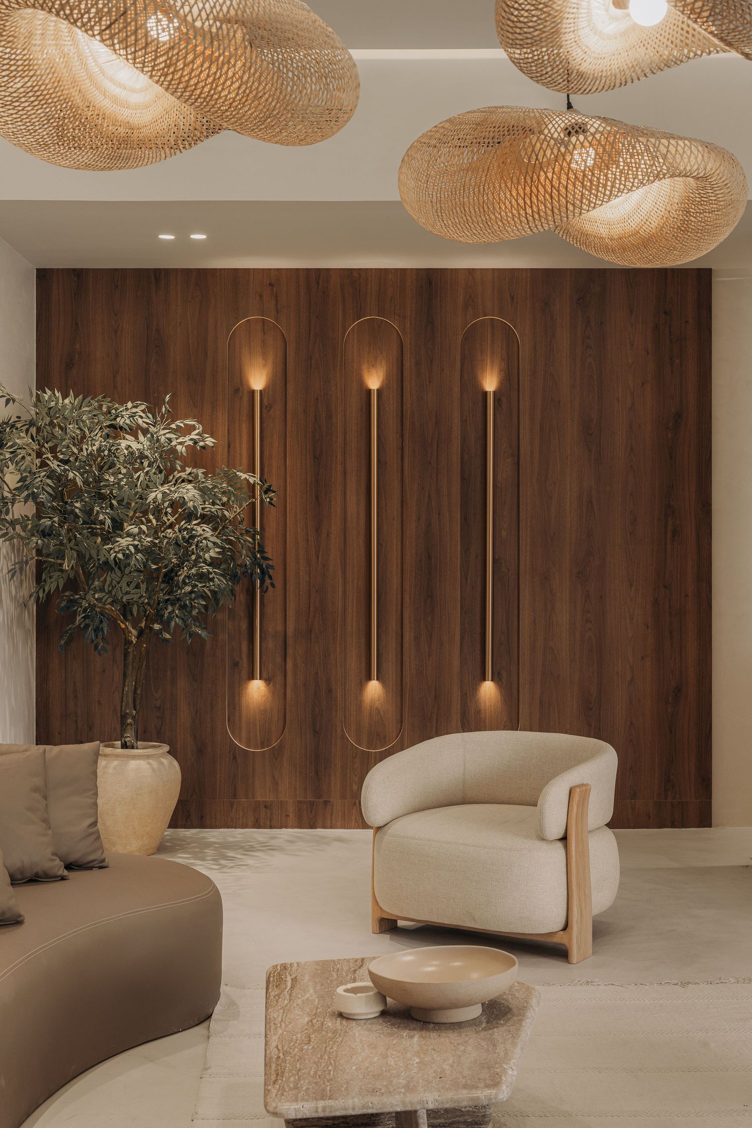 Elegant lounge with woven pendant lights, dark wood accent wall, gold sconces, neutral armchair, and indoor olive tree.