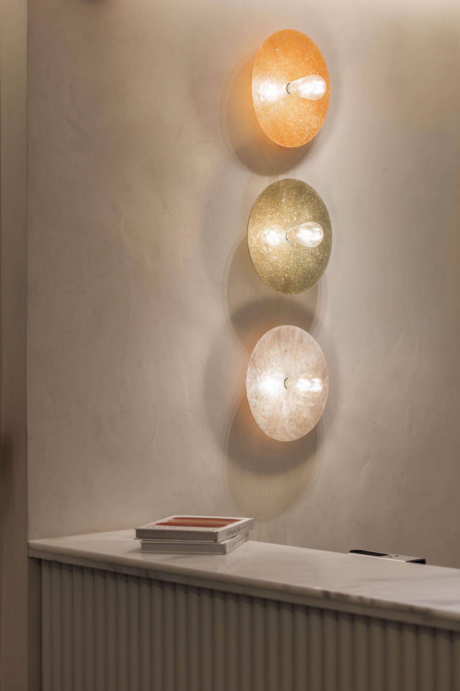 Three circular wall sconces in orange, green, and white tones mounted vertically on a beige wall above a marble counter.