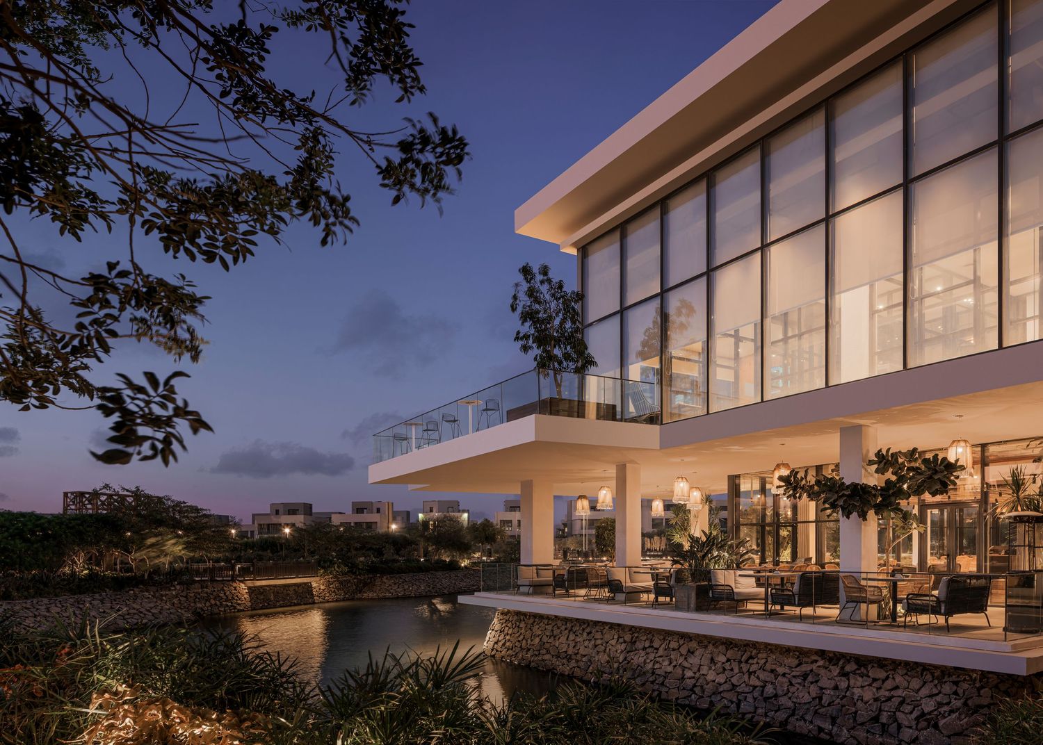 Modern luxury villa with floor-to-ceiling windows illuminated at dusk, overlooking a serene waterfront with lush landscaping.