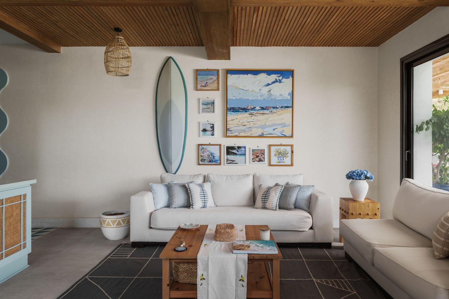 Beach-themed living room with surfboard decor, coastal artwork, white sofas, and wood ceiling.